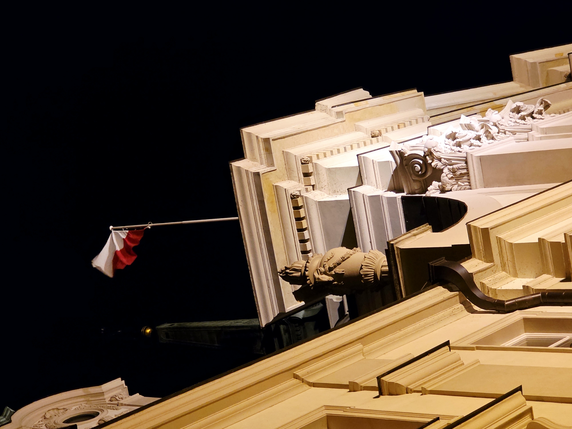 The image shows the flag of Poland, a horizontal bicolour of white and red, flying from a flagpole. The flag is positioned against a dark night sky, with the upper portion of an ornate building visible in the foreground. The building's facade is illuminated, revealing architectural details such as columns, cornices, and decorative carvings. A pointed spire, likely part of a church or tower, is visible behind the flag. The overall impression is of a historic building illuminated at night, with the national flag prominently displayed. The scene is likely located in Warsaw, Poland, given the presence of the Polish flag and the architectural style.
