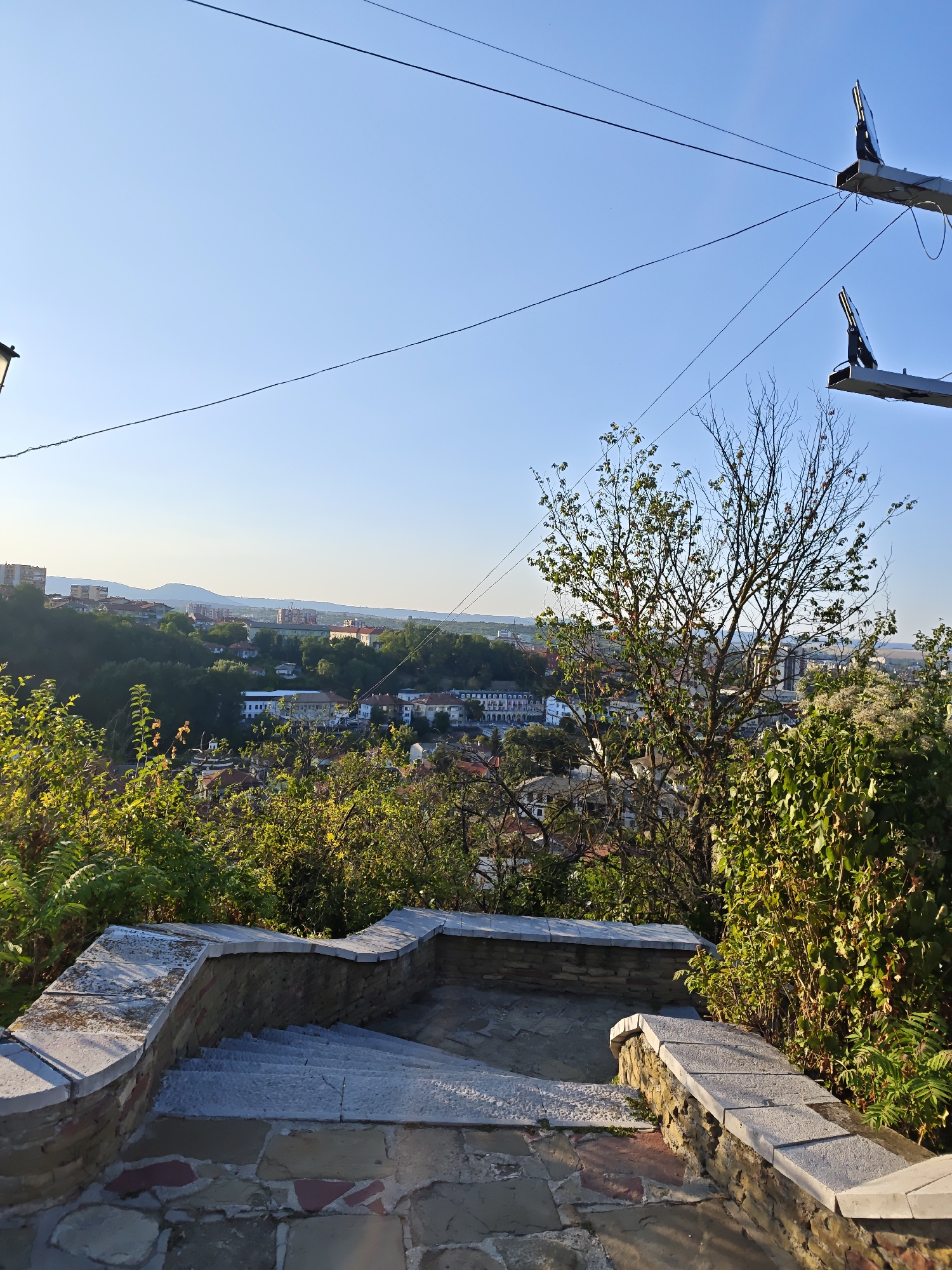 The image depicts an outdoor scene overlooking the city of Lovech, Bulgaria. In the foreground, a stone staircase with a rustic stone wall curves to the right. Sunlight illuminates the stairs and the wall. Beyond the stairs, lush green foliage and a bare-branched tree with some remaining leaves fill the middle ground. The background reveals a cityscape with buildings nestled among rolling green hills. The sky is a clear, bright blue. Two utility poles with wires are visible in the upper portion of the frame. The overall impression is a panoramic view of a historic European city on a sunny day.