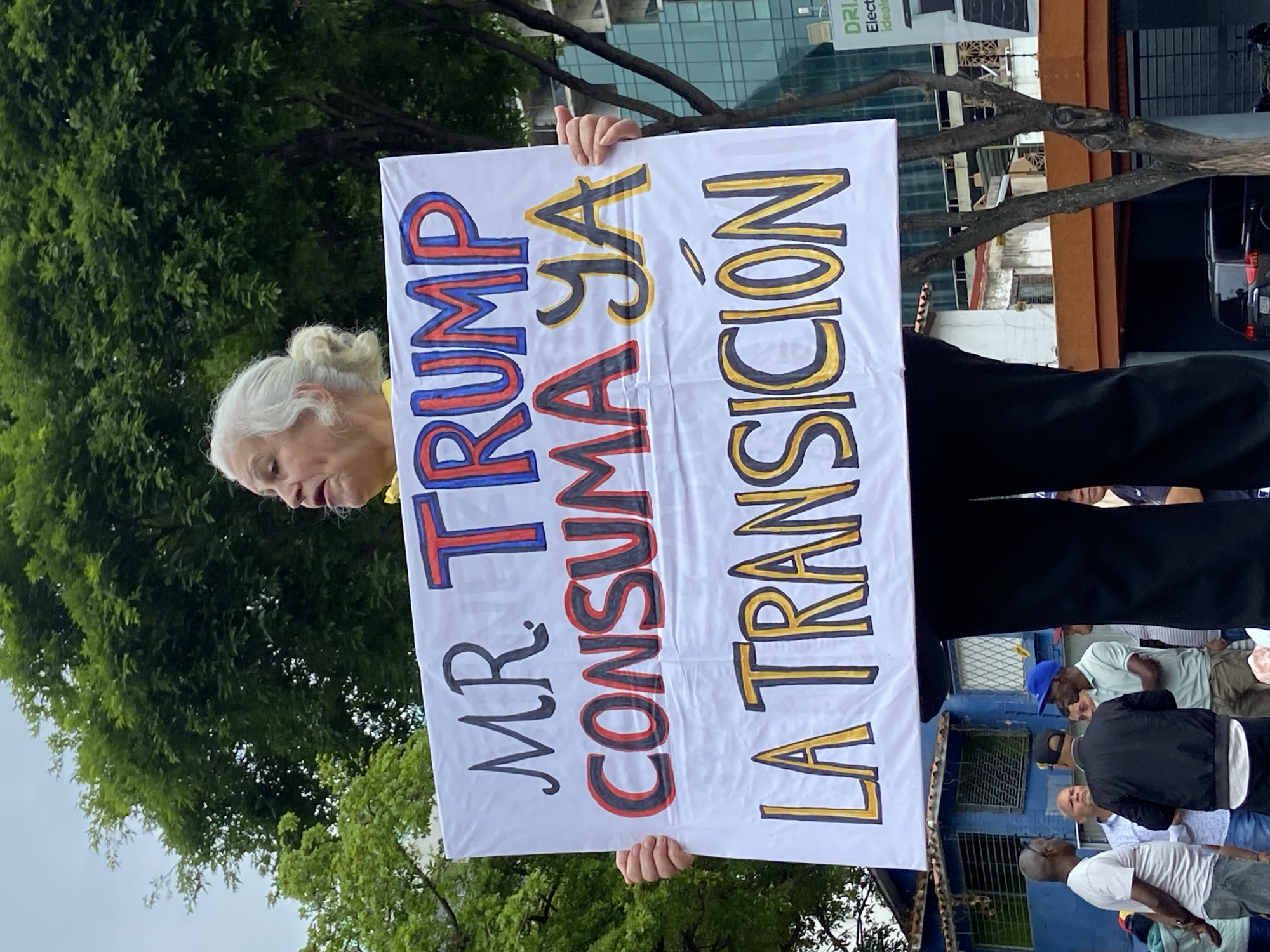 The image features an elderly person, likely a woman, with white hair pulled back, holding a large protest sign. She wears a yellow garment under dark clothing. The sign is white with large, hand-drawn letters in various colors. The text reads: "MR. TRUMP CONSUMA YA LA TRANSICIÓN." The words "MR. TRUMP" are outlined in blue and red. "CONSUMA YA" is outlined in red and black. "LA TRANSICIÓN" is outlined in yellow and black. The English translation is "Mr. Trump, consummate the transition now."
The scene is an outdoor, urban environment during the daytime. Lush green trees fill the background on the left and top. To the right, behind the person and sign, are urban buildings with visible windows and architectural elements, along with a partial view of a dark vehicle. Several other people, mostly men, are partially visible in the lower right background, suggesting a public gathering or demonstration. The person holding the sign appears focused, looking towards the left side of the frame. The overall atmosphere suggests a political event or protest taking place in a city setting, consistent with Caracas, Venezuela.