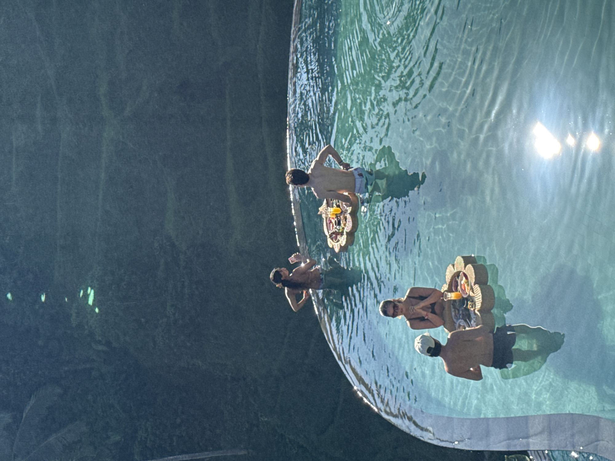 A bright, aerial view captures four individuals enjoying an infinity pool that overlooks a vast, lush green landscape, typical of Tegallalang, Indonesia. The image is oriented as if looking down onto the scene, with the pool forming a horizontal edge at the bottom of the frame, beyond which the dark, dense foliage of the jungle or rice terraces extends.
Four people are partially submerged in the clear, turquoise water of the pool. On the far left, a man with dark hair is visible from the chest up, holding a beverage. To his right, another man, also shirtless and with dark hair, stands with his back mostly to the viewer, next to one of two elaborate floating wicker trays. In the foreground on the right, a woman with light hair and sunglasses faces slightly towards the camera, positioned around a second, larger floating tray. Beside her, a man wearing a white cap is also partially submerged, facing away from the camera, near the same tray.
The floating trays are well-stocked, with the more prominent one holding glasses of orange juice, other drinks, and various food items, suggesting a floating breakfast or refreshment service. Strong sunlight creates bright glares and ripples on the water's surface, indicating a clear, sunny day, likely in the morning or late afternoon. The scene conveys a sense of serene leisure and enjoyment in a luxurious tropical setting. No visible text is present in the image.