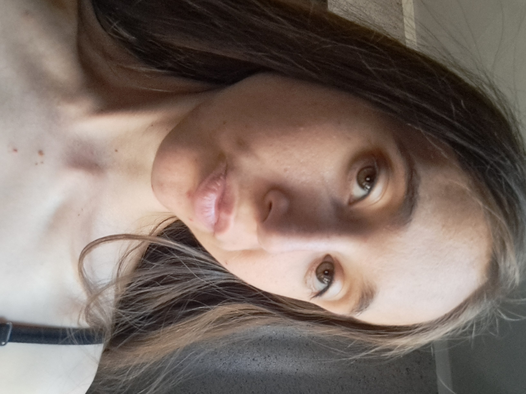 The provided image is a close-up selfie of a young adult female with long, brown hair. She is looking directly at the camera with a slightly puckered expression. Her skin appears fair with some minor blemishes and freckles visible on her face and décolletage. She is wearing a black bra strap on her right shoulder. The background is out of focus and appears to be an indoor setting with a textured wall. There is no indication of the city of Vypasne, Ukraine, or any other specific location in the image.