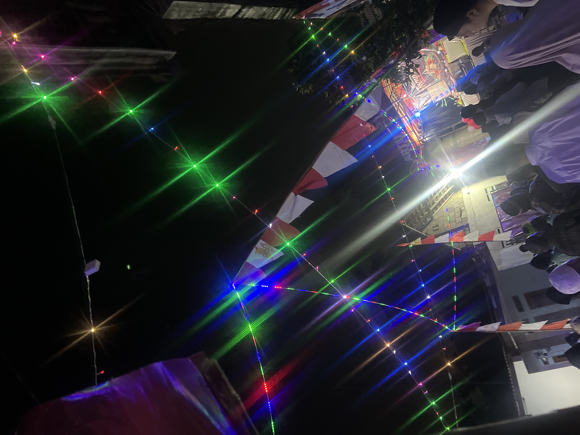 The image depicts an outdoor night scene in Klapanunggal, Indonesia, likely during a celebratory event. A string of colorful LED lights, red, green, blue, and pink, are strung across the dark sky, creating a festive atmosphere. Several Indonesian flags, red and white, are visible, some hanging loosely and others adorning structures. In the background, a stage is set up with bright lighting and what appears to be a banner or screen displaying graphics. A crowd of people, mostly men wearing peci or caps, are gathered in front of the stage, some are seated, others are standing. The lighting from the stage and the string lights illuminates the gathering. The environment suggests a local community event, possibly a festival or performance, taking place in an urban or semi-urban area of Klapanunggal.