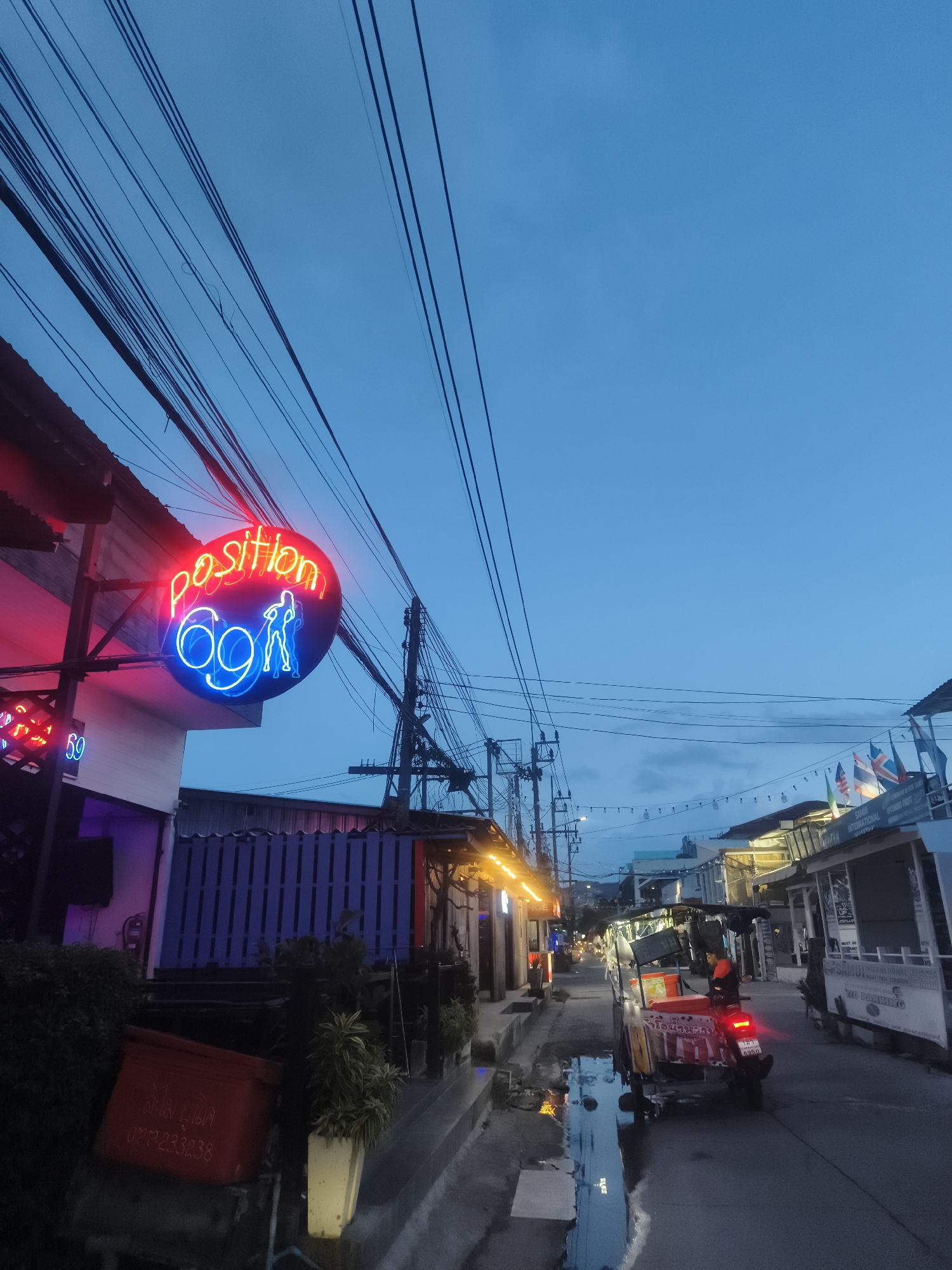 The image shows a street scene in Baan Chaweng, Thailand, at dusk. Prominently displayed is a neon sign reading "position 69" with a suggestive graphic, indicating an adult entertainment establishment. Power lines crisscross above the street. The street itself is narrow and appears wet, reflecting the surrounding lights. A three-wheeled vehicle, possibly a food cart, with Thai writing on it, is parked on the right side of the street. Various commercial establishments line both sides of the street, some with visible signage and flags. A red plastic bin is visible near the curb on the left.
