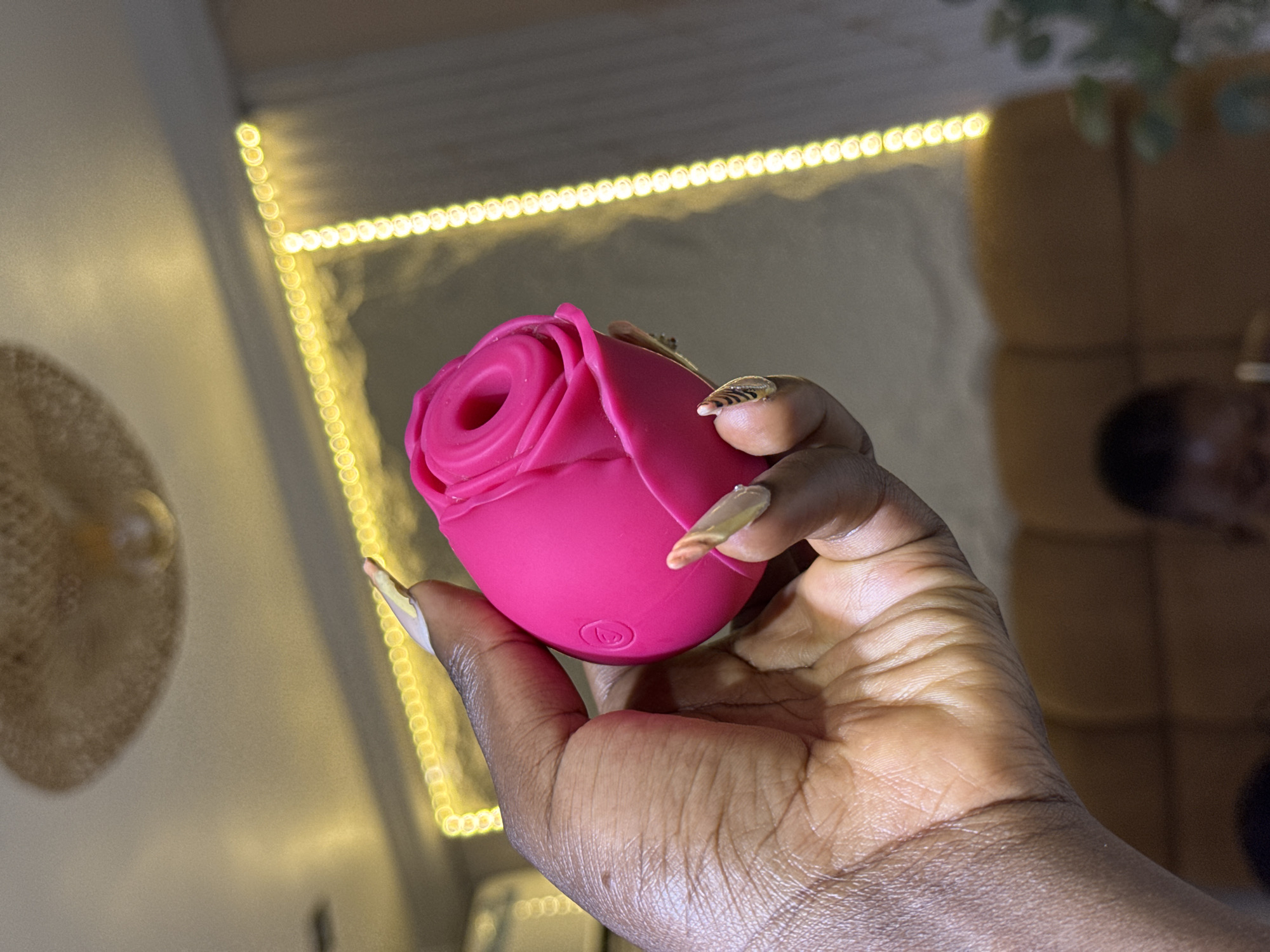 A close-up, high-angle shot shows a person's hand, with dark skin and long, light-colored nails, holding a bright pink rose-shaped object. The object appears to be made of silicone and has a textured surface that mimics the petals of a rose. The person's thumb is positioned underneath the object, while their index and middle fingers gently cradle its side. The hand is in focus, with the object also sharp.
In the background, a string of warm yellow LED lights is visible, arranged in a rectangular pattern along what appears to be a wall or shelf. The lights are out of focus and create a soft glow. To the left of the hand, a circular, woven object, possibly a decorative piece or a basket, is partially visible and also out of focus. Further in the background, a blurred reflection in a mirror or glossy surface shows a glimpse of a brown textured area, possibly furniture, and a hint of green foliage from a plant. The overall lighting suggests an indoor setting, possibly during the evening or in a dimly lit room, with the artificial lights providing illumination and ambiance.
There is a small, circular logo on the rose-shaped object, though the details are not clear enough to discern any text. The person's nails have a design with tiger-stripe patterns. The scene evokes a sense of intimacy and personal care, possibly related to self-care or beauty products. The location is stated as Abuja, Nigeria.