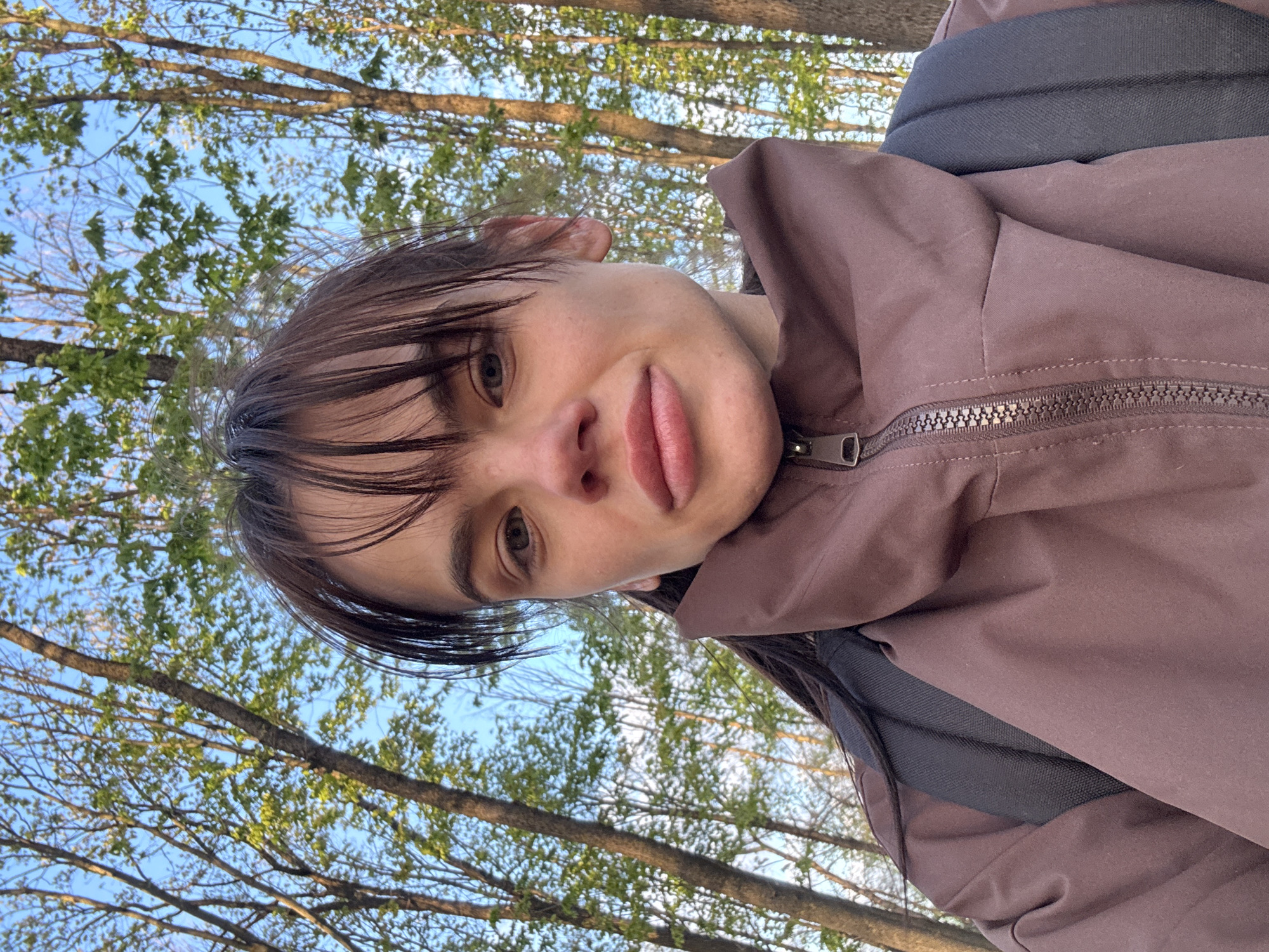 A selfie of a person with bangs is taken outdoors with tree branches and a blue sky visible in the background. The person is wearing a brown jacket with a zipper. A black backpack strap is visible over the jacket. No specific location in Rostov-on-Don is discernible from the image.