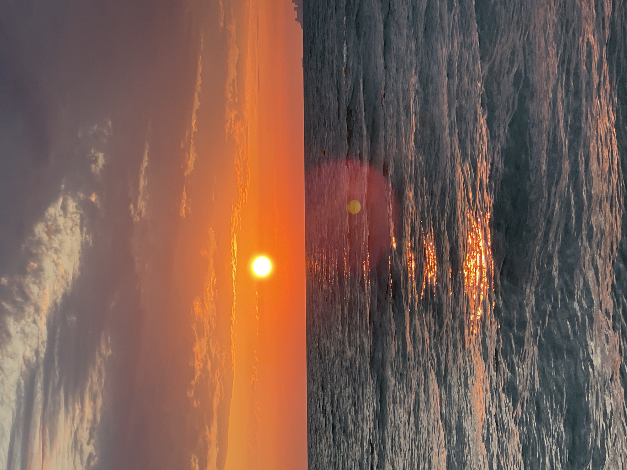 The image captures a sunset over the Black Sea, likely near Khostinskiy, Russia, given the visible cityscape on the horizon. The sun, a bright orb, is positioned low in the sky, casting a vibrant orange and red glow across the horizon and the sky above. Wispy clouds, tinged with pink and purple, are scattered across the sky. The sea is a deep blue-grey, with gentle waves reflecting the warm hues of the sunset. A narrow path of shimmering sunlight stretches across the water towards the viewer. In the distance, a cluster of buildings forming a city skyline is faintly visible on the right side of the horizon. No people or specific objects are identifiable in the foreground or mid-ground of the scene.