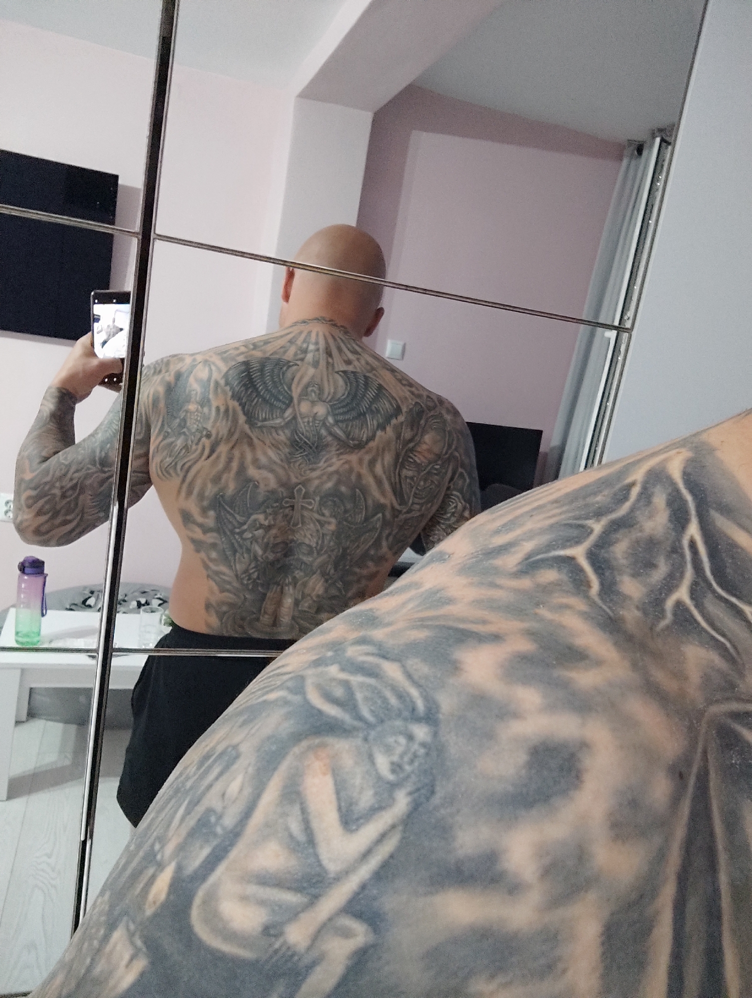 The image displays a heavily tattooed man taking a mirror selfie indoors. The man, who is bald and seen from the back, stands before a large mirrored surface, likely a wardrobe, using a smartphone in his right hand to capture his reflection. The phone's screen partially shows the room.
His entire back is adorned with extensive, dark-toned tattoos. The upper back features a prominent winged figure, possibly an angel or demon, from which rays emanate. Below this, another central design shows a horned, demonic-like figure with a cross on its head, surrounded by intricate patterns and other figures. Both of his arms are also fully tattooed with dark, flowing, and abstract designs, including a reclining figure visible on his right upper arm in the foreground.
The setting is a room with light-colored, possibly pale pink, walls and a light-toned floor. Reflected in the mirror are various room elements: a dark rectangular screen (television or monitor) to the left, a white table with a glass top, and a purple and green water bottle. White vertical blinds or curtains are visible on the right side of the reflection, along with a white electrical switch or outlet on the wall. The scene is brightly lit, suggesting daytime. The location is Burgas, Bulgaria. No visible text is present in the image.