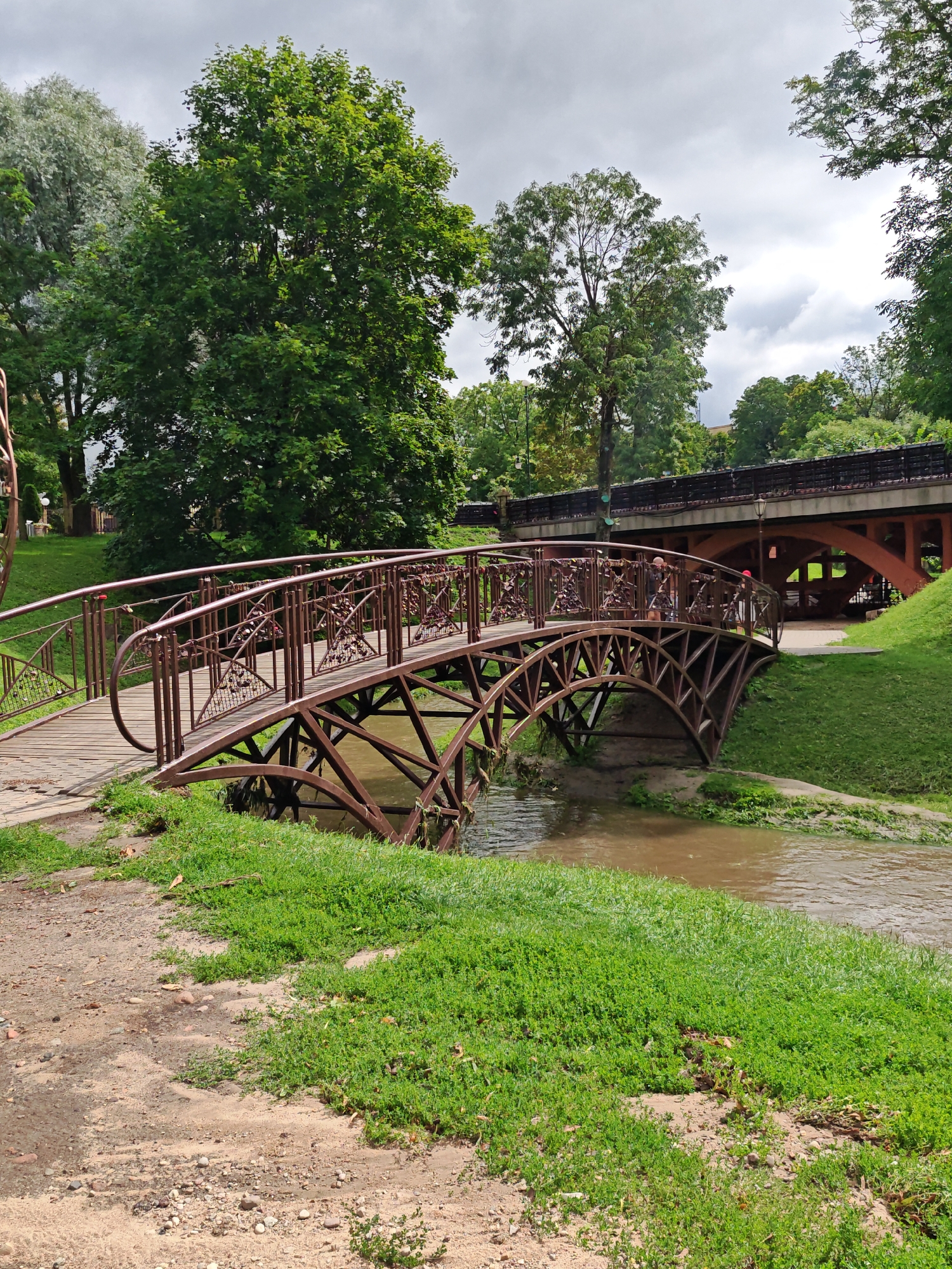 A small decorative bridge arches over a narrow, muddy stream in a park setting, likely in Hrodna, Belarus. The bridge is constructed of dark brown metal with intricate scrollwork on the railings and decorative triangular patterns on its sides. Padlocks are attached to the railings, suggesting it is a romantic landmark. The bridge connects to a paved pathway on the left and leads to a grassy bank on the right. In the background, a larger, more utilitarian bridge with an arched structure is visible, also crossing the stream. Lush green grass covers the ground, and mature trees with dense foliage dominate the background. The sky is overcast, with a mix of grey clouds and brighter patches. Two individuals are present on the decorative bridge, one appearing to be looking over the railing and the other closer to the center of the bridge. The overall scene depicts a tranquil park environment with elements of natural beauty and human adornment.