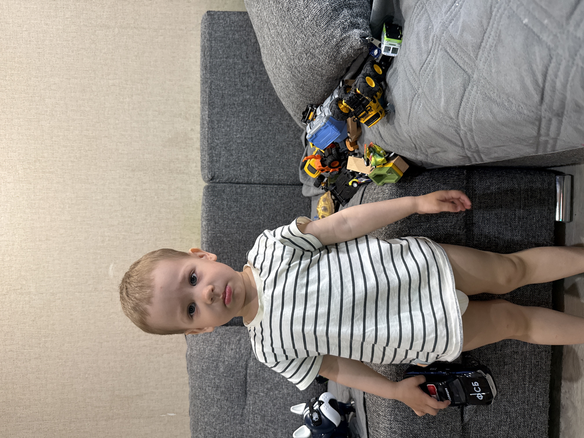 A young boy is standing on a gray couch with a variety of toy vehicles piled next to him. He's wearing a black and white striped shirt and holding a black and blue toy car. The room has beige-colored walls. It's impossible to determine the location as Rusanovo, Russia with only the given image.