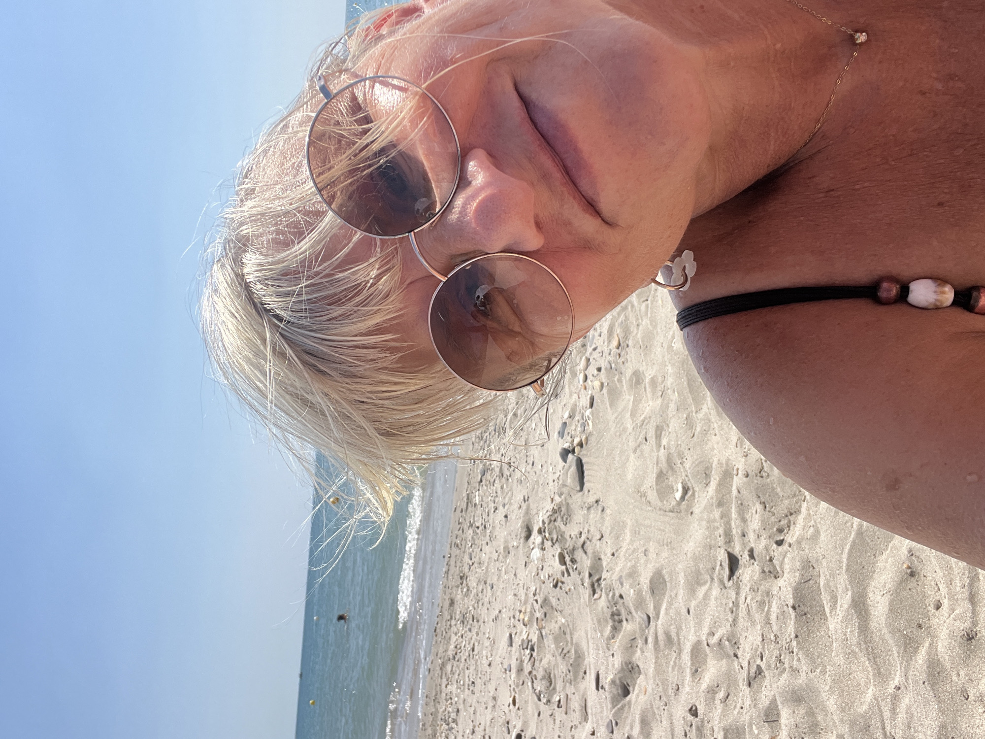 A woman with blonde hair and sunglasses is taking a selfie on a sandy beach. The sea is in the background, with a clear blue sky above. The woman is wearing a black bikini top and a gold necklace. She appears to be smiling and relaxed. The location is the beach in Villeneuve-lès-Maguelone, France.