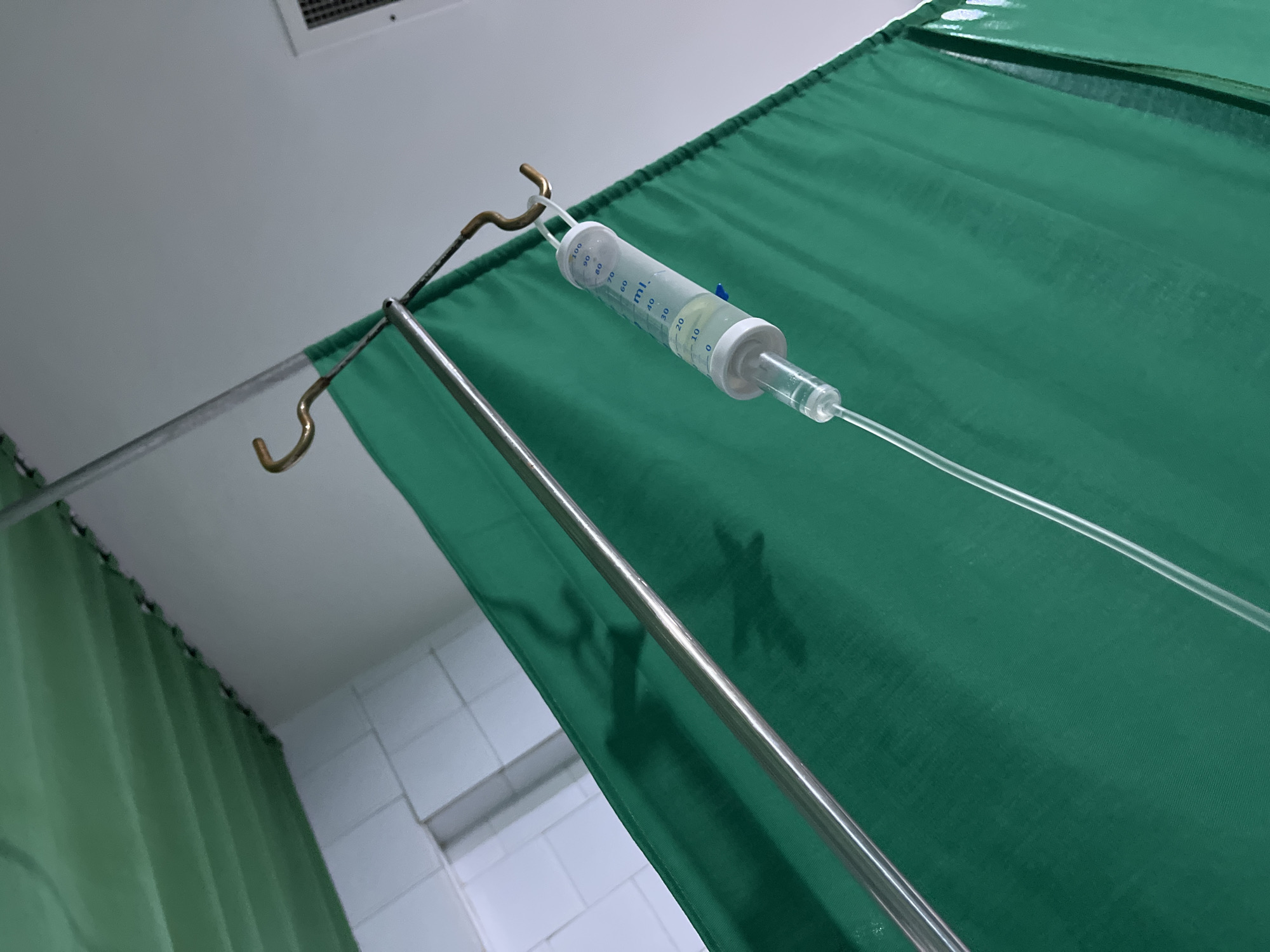 The image shows a medical IV bag hanging from a metal stand, with a green curtain in the background. The IV bag is partially filled with fluid. The scene appears to be in a hospital or clinic, given the presence of the IV and curtain. The location in Juan Griego, Venezuela is not directly identifiable from the image alone.