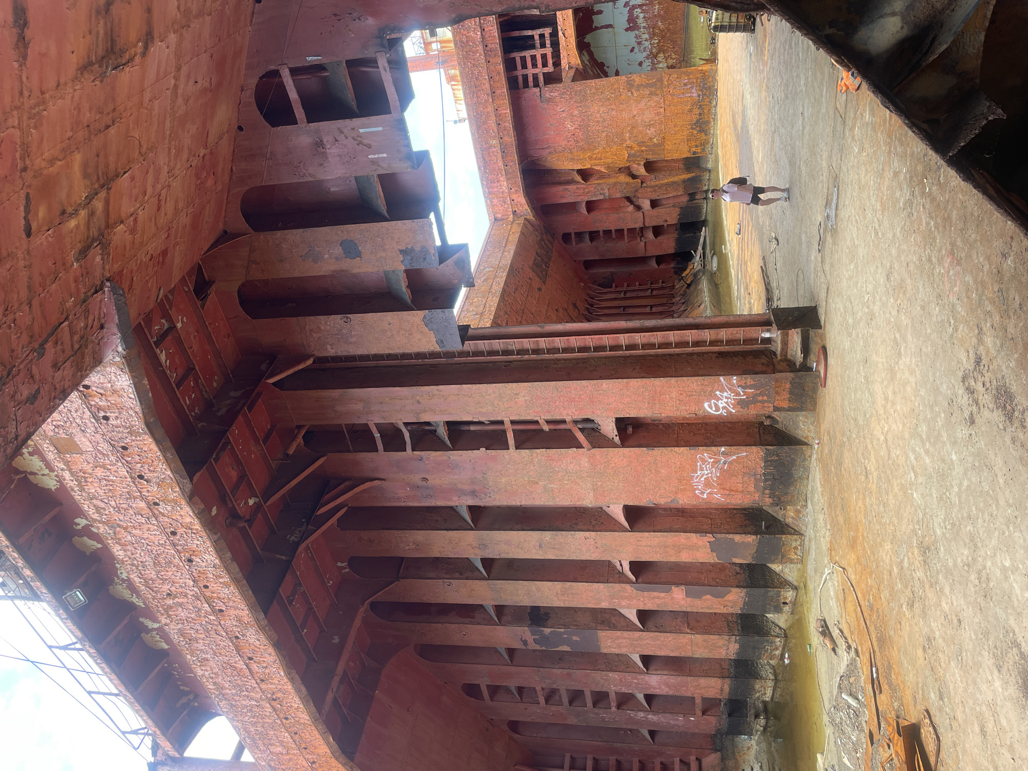 The image depicts the interior of a large, derelict ship, likely a cargo vessel, in Kabardinka, Russia. The hull is heavily corroded, with exposed metal and peeling red paint evident throughout. Large, vertical structural supports dominate the scene, creating a cavernous interior. A person, wearing a pink backpack and dark shorts, stands in the open area of the ship's interior, facing away from the camera. The ground is covered in sand and debris. Water is visible in the lower left portion of the image, suggesting the ship may be partially submerged or in a dry dock. The sky is visible through openings in the upper structure.