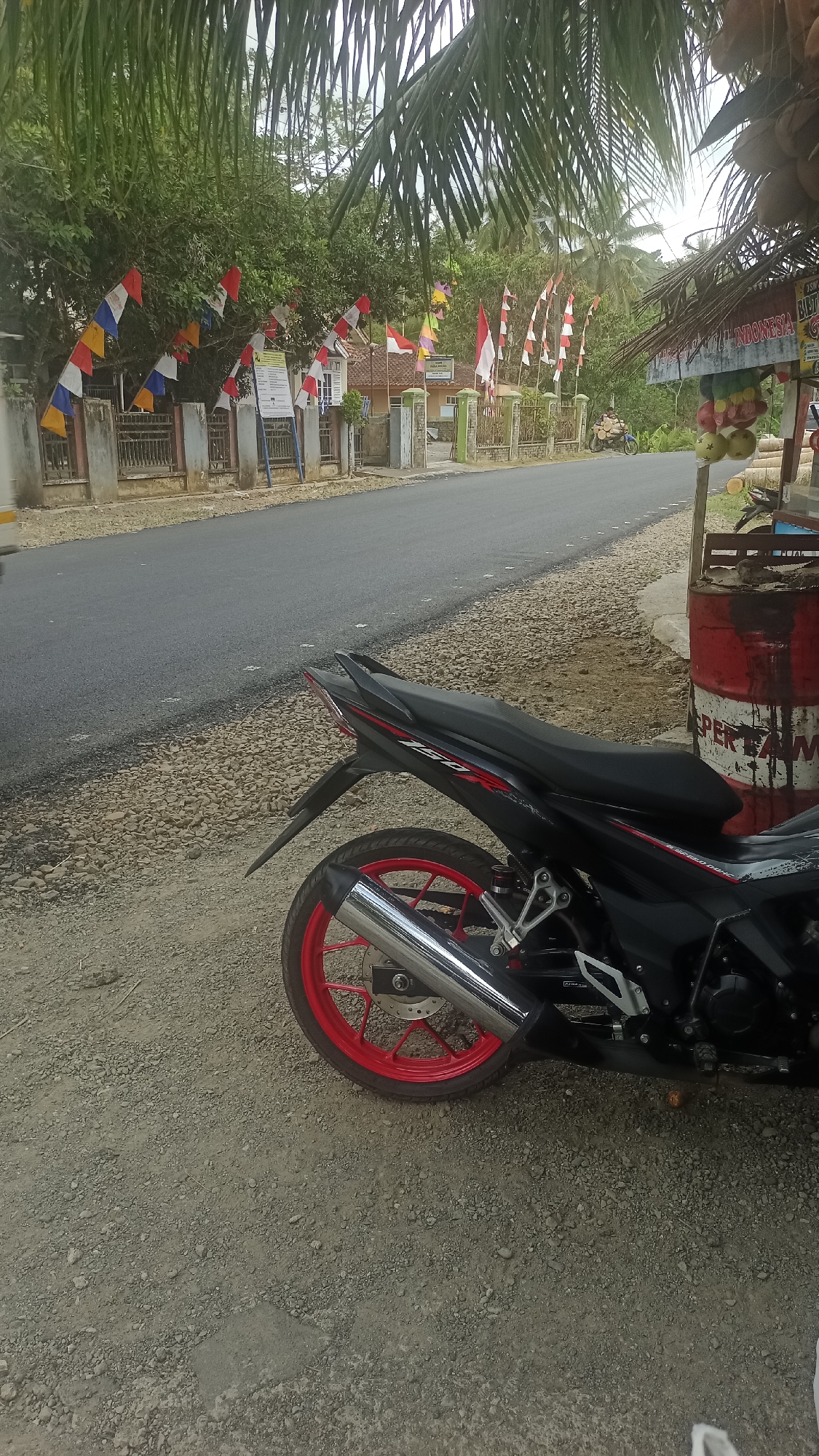 In Cibeber, Indonesia, a black motorcycle with red accents is parked on the gravel shoulder of a paved road. The motorcycle's rear wheel, featuring a red rim and a silver exhaust pipe, is prominently visible. The road is lined with trees and buildings adorned with Indonesian flags, suggesting a national holiday or celebration. In the background, a small roadside stall displays fruits and a red barrel. The overall scene depicts a quiet, possibly rural or suburban, street environment.