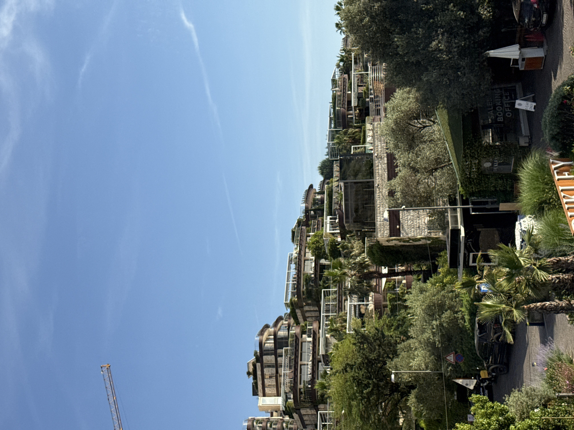 The image depicts a view of Budva, Montenegro, from a high vantage point. The foreground is dominated by buildings and greenery, including palm trees and other vegetation. The buildings are multi-storied with balconies and a modern architectural style. There is a “Sales Booking Office” visible. In the background, there's a clear blue sky with faint cloud formations. A construction crane is situated on the left side of the image, reaching upwards into the sky. The environment is a mix of urban development and natural landscape.