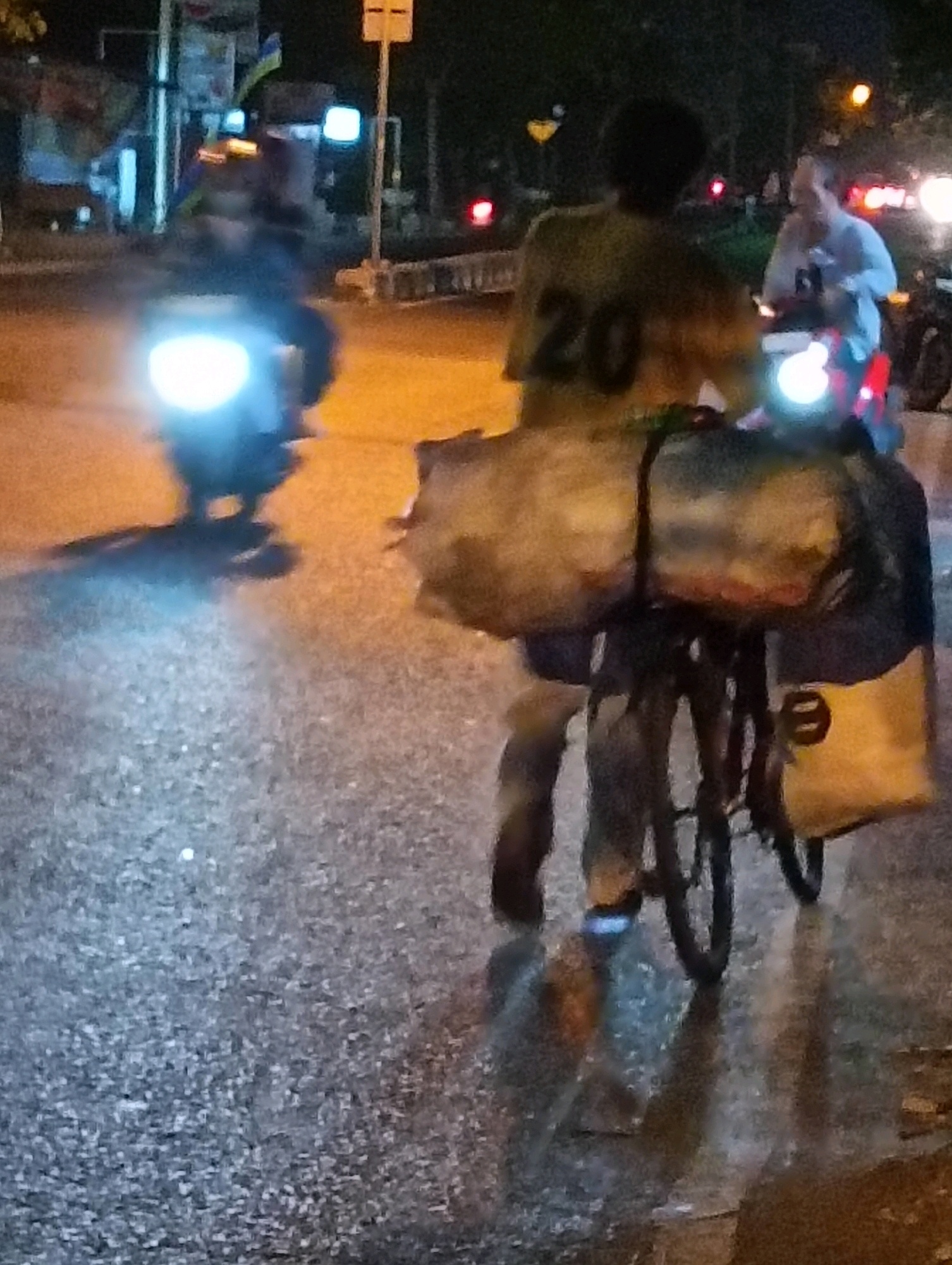At night, on a wet asphalt road in Purworejo, Indonesia, a person is riding a bicycle laden with goods. The cyclist, wearing a yellow shirt with the number "20" on the back, is partially obscured by large bags secured to the bicycle. The road surface reflects the ambient light from streetlights and vehicle headlights. In the background, another person is riding a motorcycle with its headlights on, and a traffic light is visible. The scene suggests movement and activity on a city street after dark.