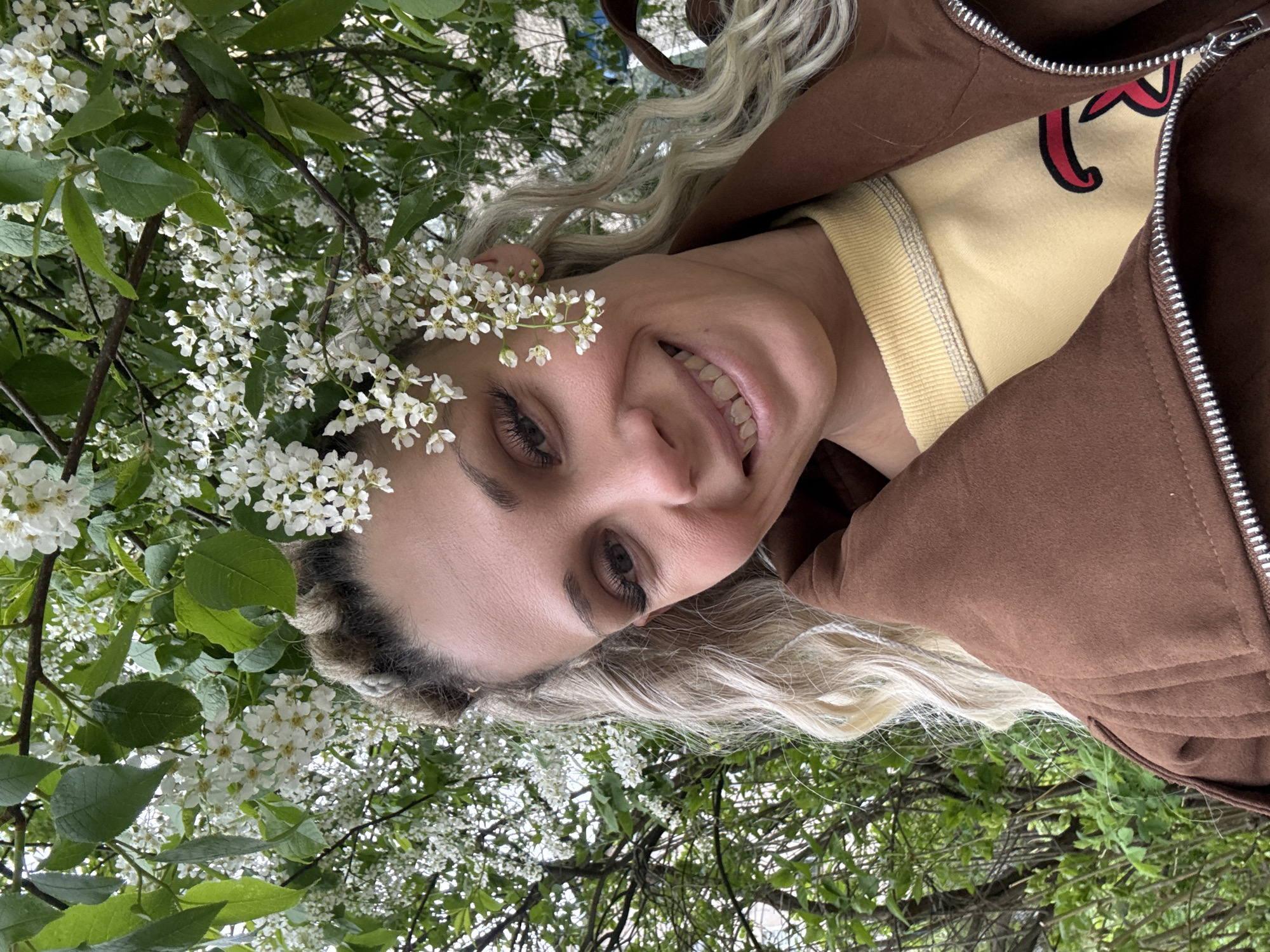 A fair-skinned woman with braided blond hair is depicted from a low angle. She is smiling and has white flowers near her face, seemingly positioned like a crown. She is wearing a brown zip-up jacket over a yellow top with a red design. The background consists of a tree with white flowers and green leaves, potentially in a park-like setting. The image was possibly taken in Rusanovo, Russia.