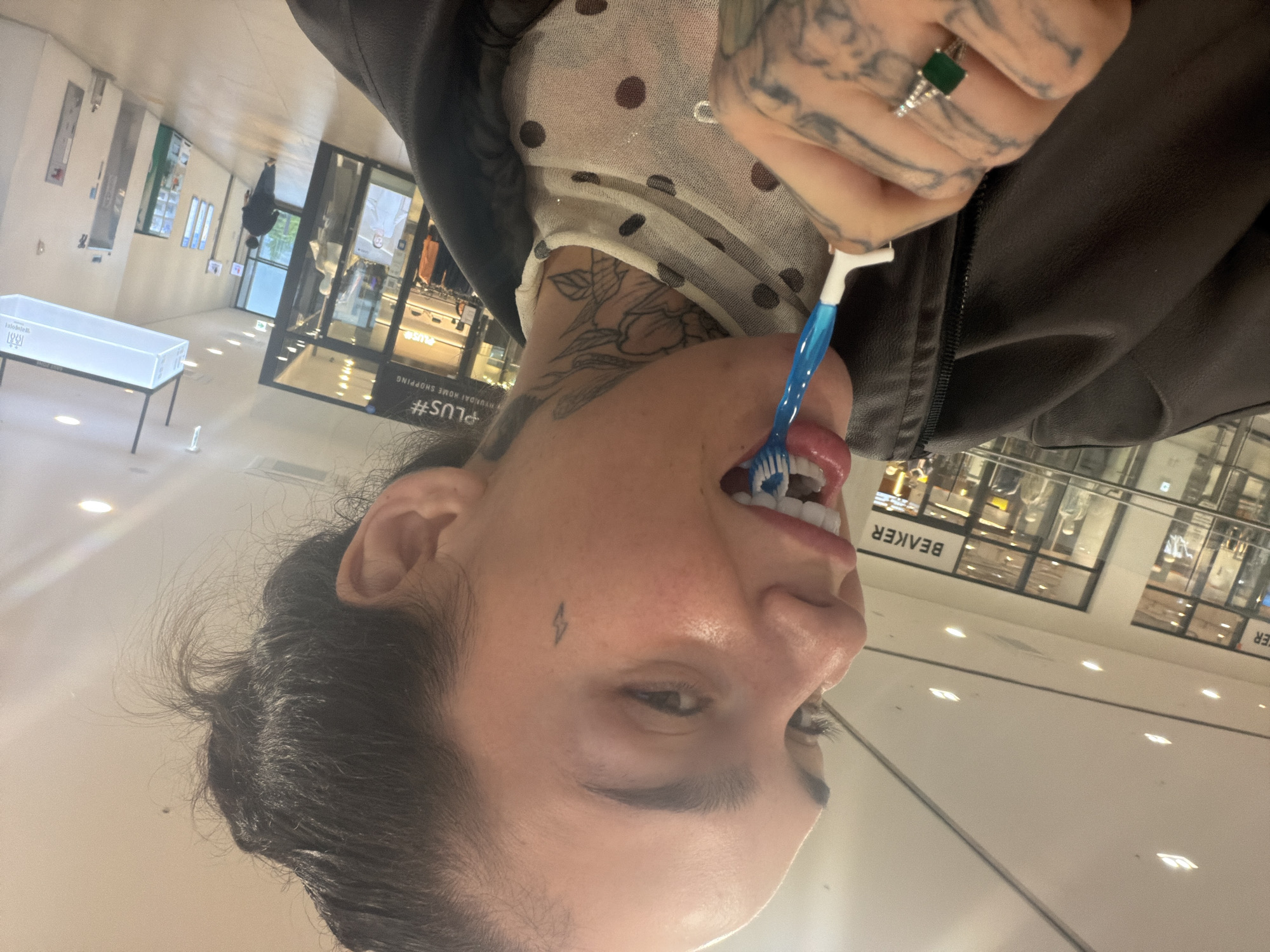 The image depicts a person brushing their teeth with a blue toothbrush in what appears to be a public space. The person has tattoos on their neck and hands, and is wearing a ring with a green stone. The background shows a retail environment with the word "BEAKER" visible, indicating a store. Based on the context, the location is Gimpo-si, South Korea.