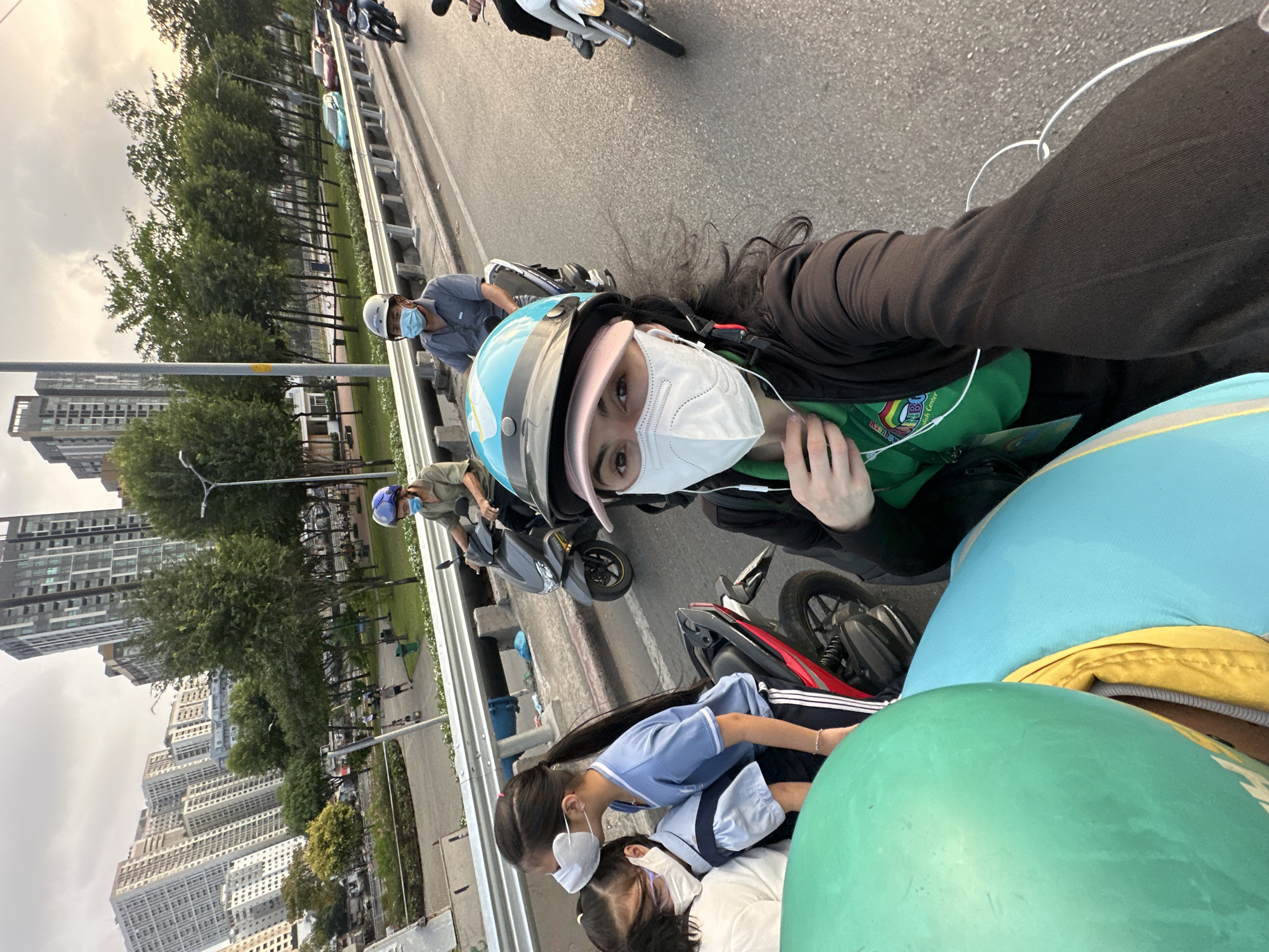 The image depicts a person wearing a blue and white motorcycle helmet, a white face mask, and earphones, seemingly taking a selfie. They are wearing a green shirt with what appears to be a logo, and a dark jacket. Other people are on the motorcycle behind them, including two children wearing school uniforms and face masks. The location appears to be a bridge or elevated road in Ho Chi Minh City, Vietnam, as evidenced by the high-rise buildings in the background, typical of the city's skyline. Other motorcyclists are present on the road.
