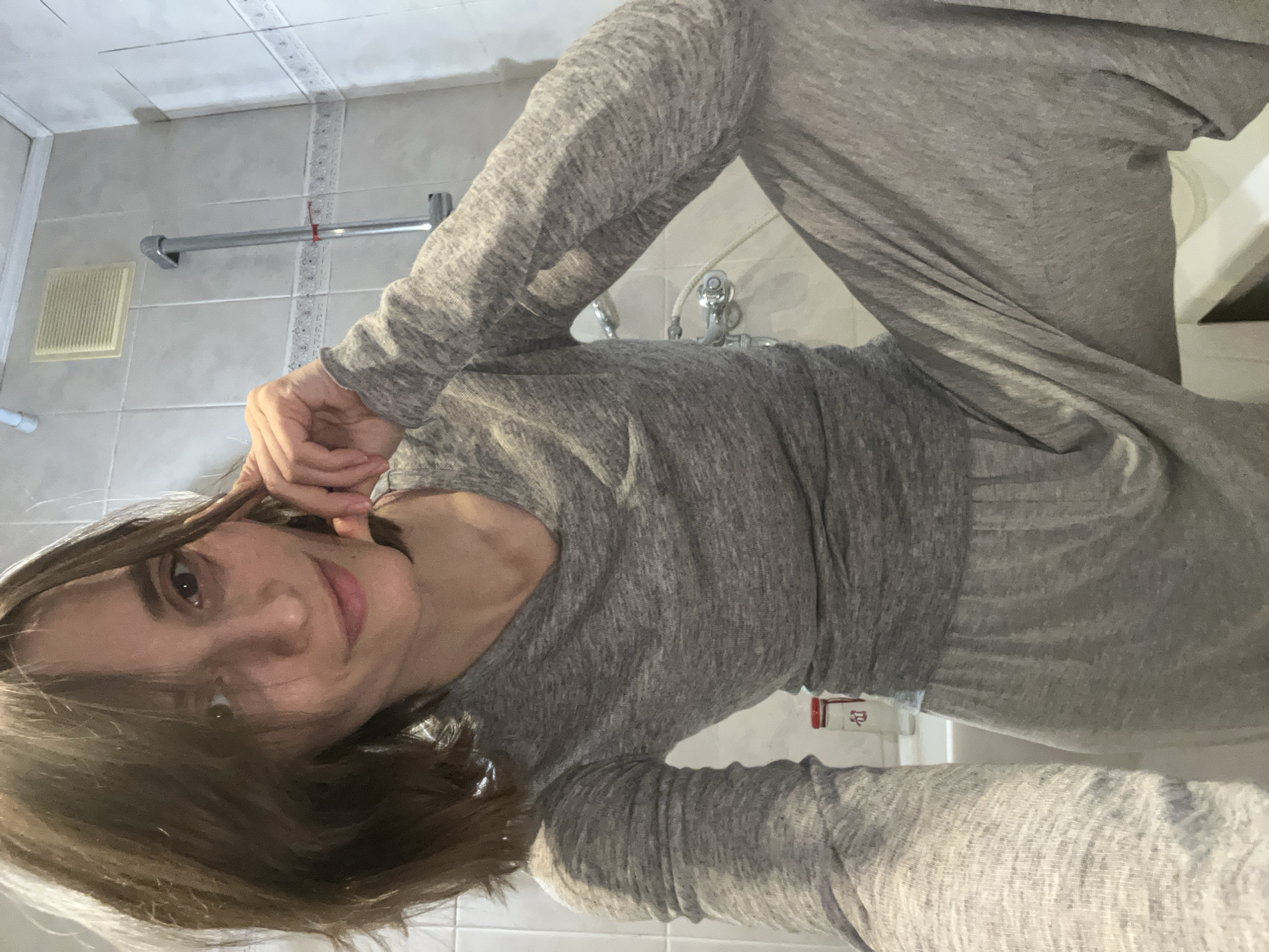 A person with brown hair and fair skin is wearing a gray long-sleeved shirt and matching pants. The individual is in a bathroom with white tiled walls and a white ceiling. Visible bathroom fixtures include a towel rack, a vent, a faucet, and a portion of a sink. The location in Moscow, Russia, is not apparent from the visual information provided.