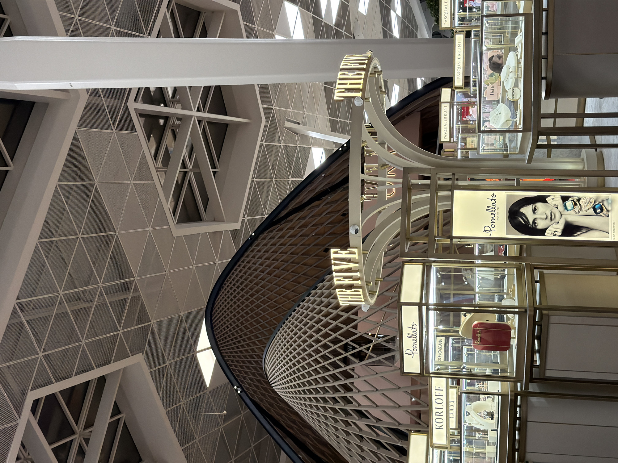The image depicts an interior view of a modern, upscale shopping area, likely a high-end mall or airport terminal, possibly in Khazar Raion, Azerbaijan. The architecture features a striking, undulating wooden structure that dominates the background, resembling a series of interconnected domes or waves. This organic form contrasts with the geometric precision of the suspended ceiling, composed of triangular panels and metal framework, illuminated by recessed lighting.
In the foreground, several retail display units are arranged, showcasing luxury jewelry. These displays are enclosed in glass cases with gold-colored frames and illuminated from above, highlighting the merchandise. Prominent signage identifies brands such as "KORLOFF GUCCI," "Pomellato," and "PASQUALEBRUNI." One display features a large advertisement or portrait of a woman adorned with jewelry. To the right, a tall, white cylindrical column rises towards the ceiling, a structural element that breaks the visual flow of the architecture. The overall atmosphere is one of luxury, sophistication, and modern design. There are no visible people engaged in any specific actions or interactions in this frame, though reflections in the display cases suggest other patrons might be present.