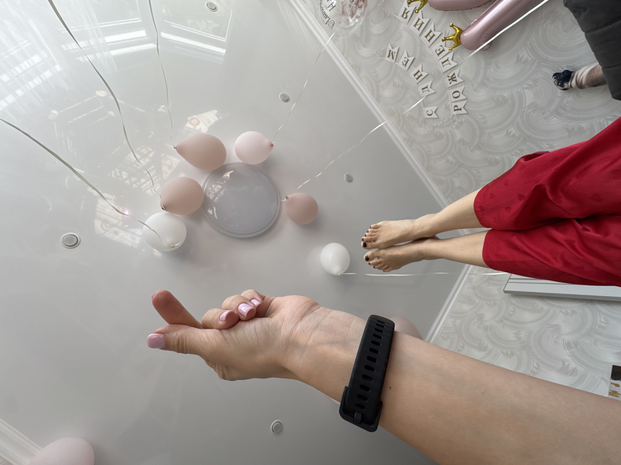 A low-angle shot looking up at a white ceiling with scattered pastel balloons, revealing a person's legs and feet in the upper right quadrant. The person wears red clothing. In the foreground, a hand with light pink nail polish extends towards the center, adorned with a black fitness tracker on the wrist. The ceiling features a circular light fixture and various other fixtures, and writing in Russian near the upper right corner indicates a birthday celebration. The location may be an apartment or interior space in Sochi, Russia based on the language and possible cultural context. The scene suggests a party or celebration viewed from a unique, ground-level perspective.