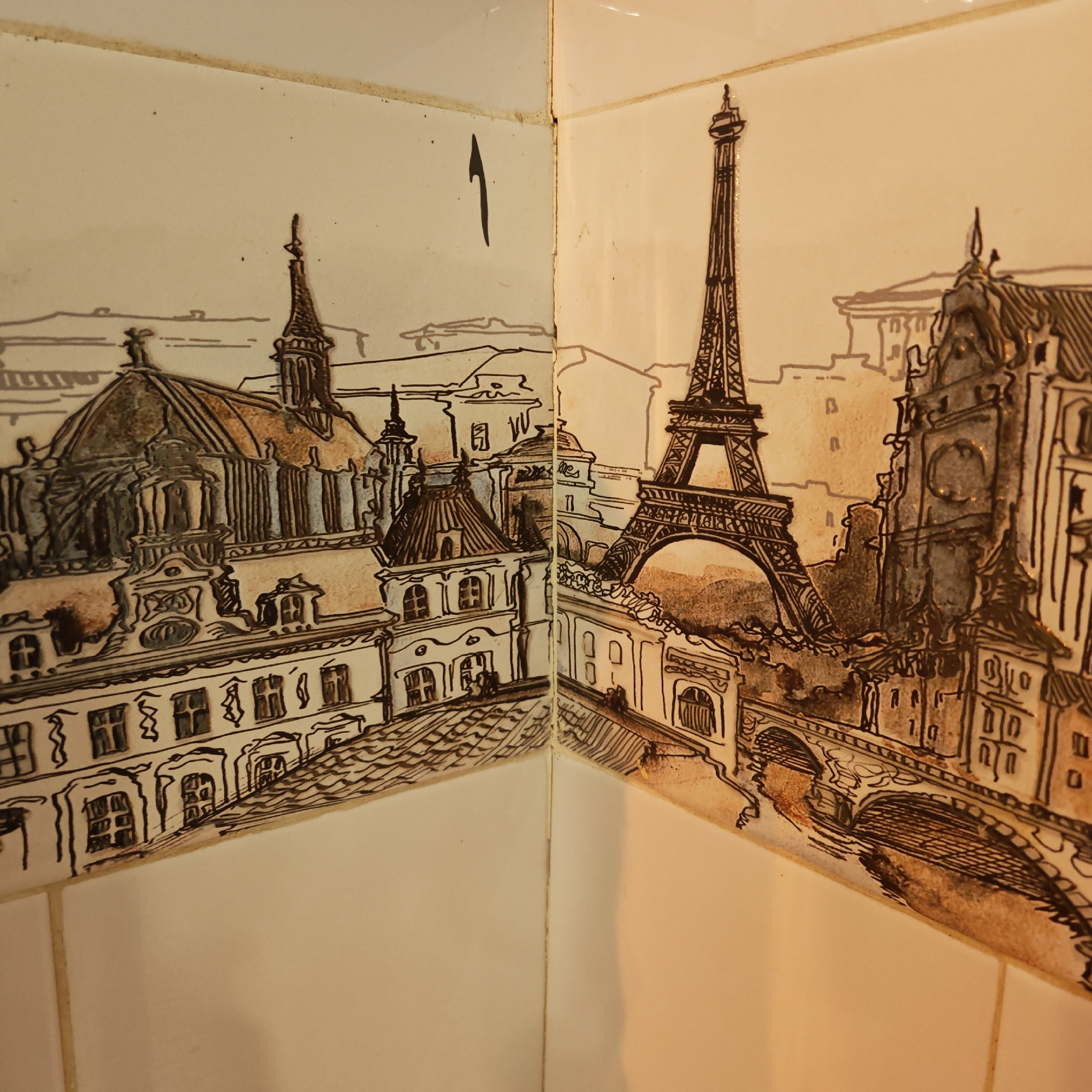 The image shows two adjacent ceramic tiles depicting a cityscape of Paris, likely a bathroom or kitchen tiling. The left tile features a building complex with a dome and towers, while the right tile displays the Eiffel Tower, buildings, and a bridge. The style resembles a sepia-toned sketch. There is no evidence or indication of Rostov, Russia.