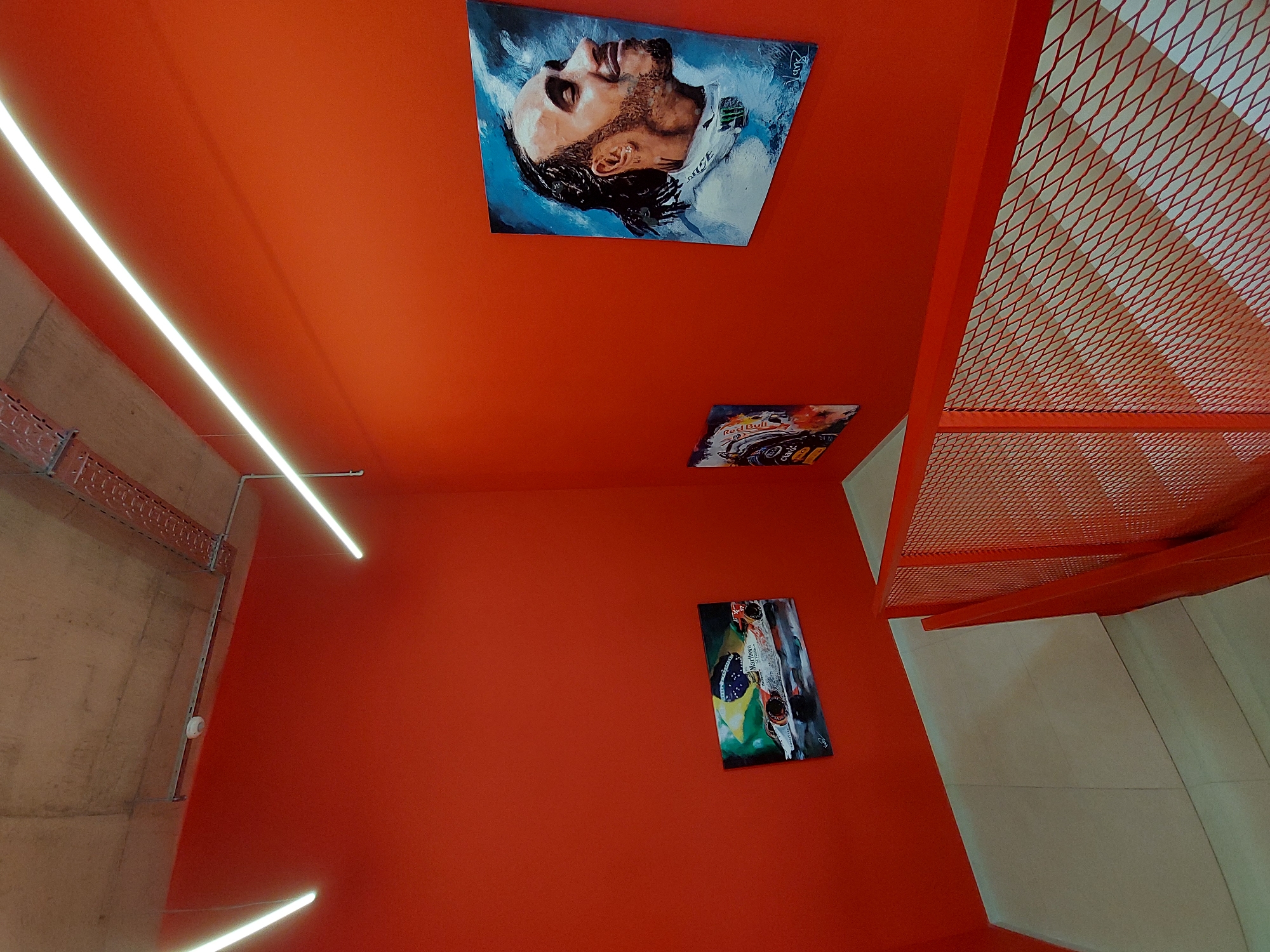 The image shows an interior space with red walls and a red ceiling. A concrete wall with conduit and a smoke detector is visible on the left. There are three paintings on the wall and one on the ceiling. The painting on the ceiling is of a man. One wall painting appears to be of a Formula 1 race car, and another depicts what looks like a Formula 1 car with a Brazilian flag and the words "Marlboro" and "Senna". To the right is a metal grate structure, likely part of a staircase or other architectural feature, and white stairs. There are two linear LED lights, one in the top left and one in the bottom left. The location and city are unknown.