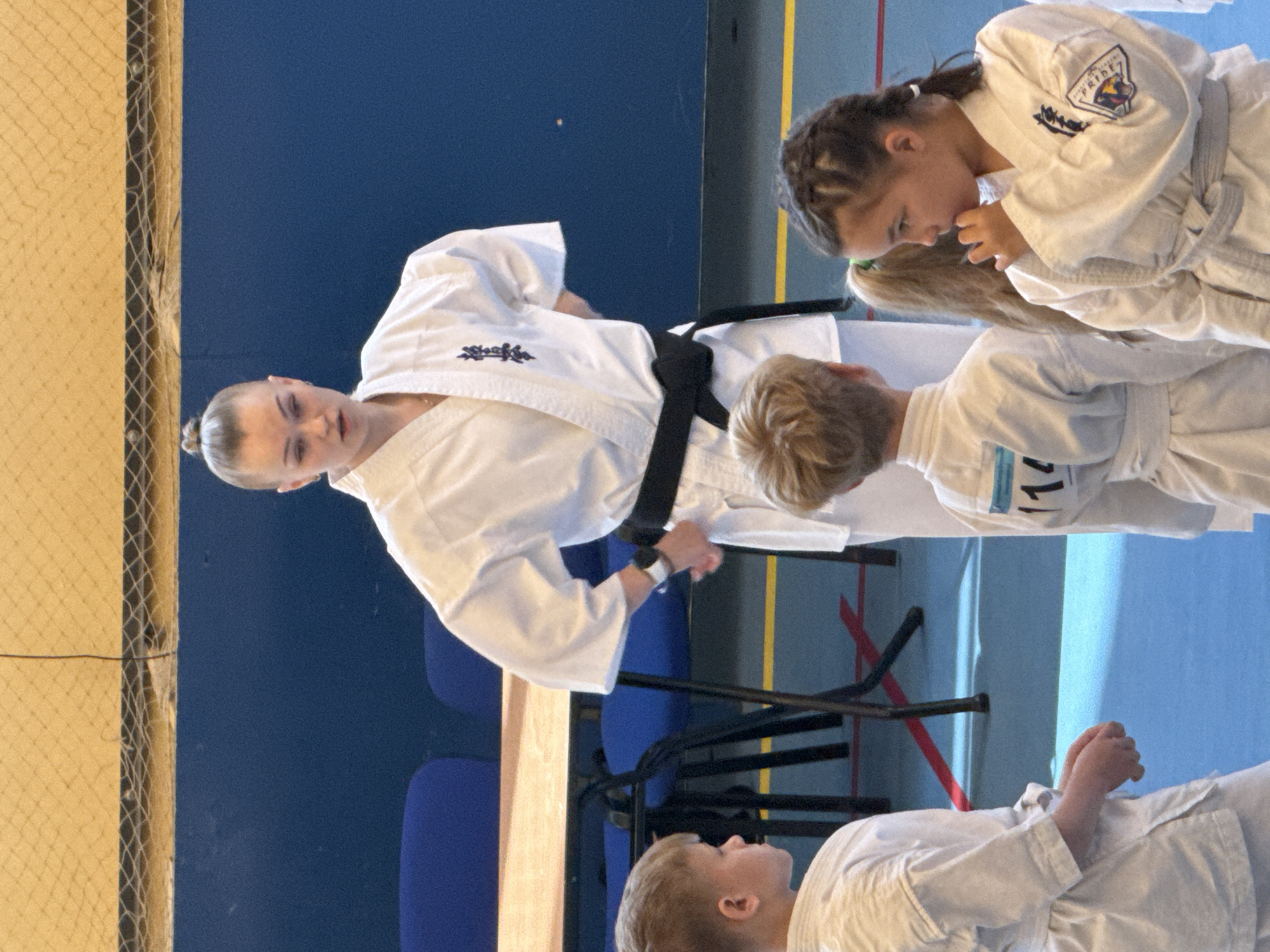 The image shows a karate competition indoors. A female karate instructor with a black belt stands overseeing three children in white uniforms. Two of the children are looking at each other, while the third is looking up at the instructor. The setting appears to be a gymnasium or sports hall. The specific location in Yekaterinburg, Russia, cannot be determined from the visual information.