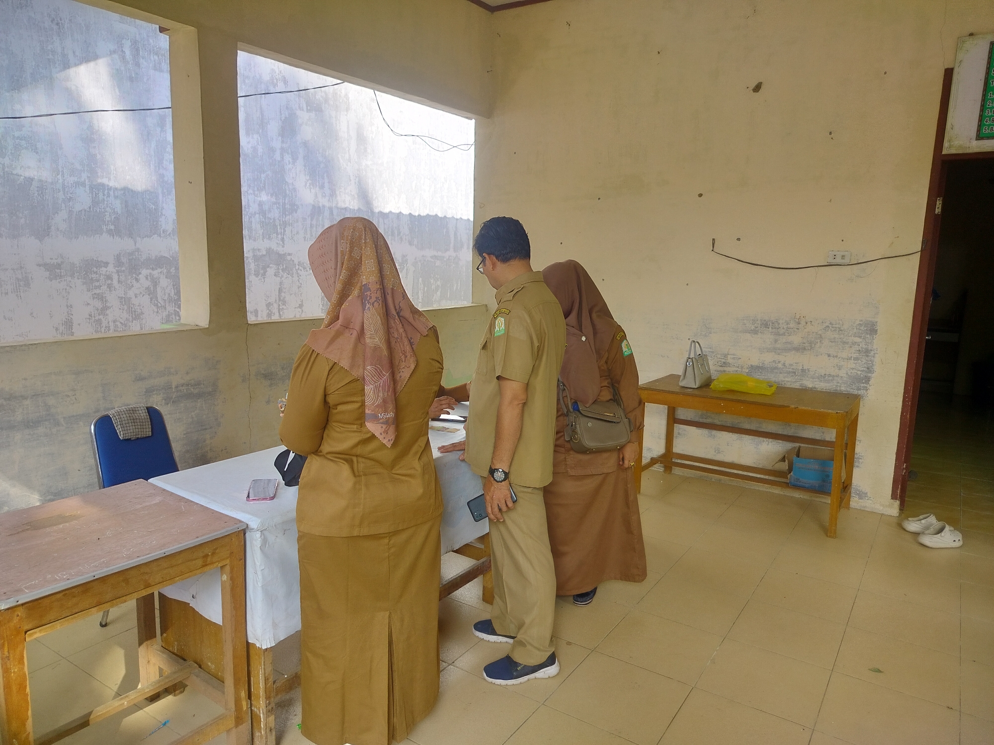 In Purwodadi, Indonesia, three individuals dressed in tan government uniforms are gathered around a table. The person in the center, a male, is wearing a short-sleeved shirt and pants, holding a smartphone in his right hand. Two females, one on the left and one on the right, are wearing long-sleeved shirts and skirts. The female on the left is partially visible from the back, while the female on the right is facing the table. A table covered with a white cloth is situated in the foreground, with papers and a smartphone on it. To the left of this table is another wooden desk. In the background, a yellow shelf unit holds a gray handbag and a yellow item. The environment appears to be an indoor space with tiled flooring and cream-colored walls. Natural light streams in from two windows on the left side, casting shadows. A blue chair with a checkered cushion is positioned near the table. Two pairs of white clogs are visible near the right wall. A doorway is present on the right side of the image.