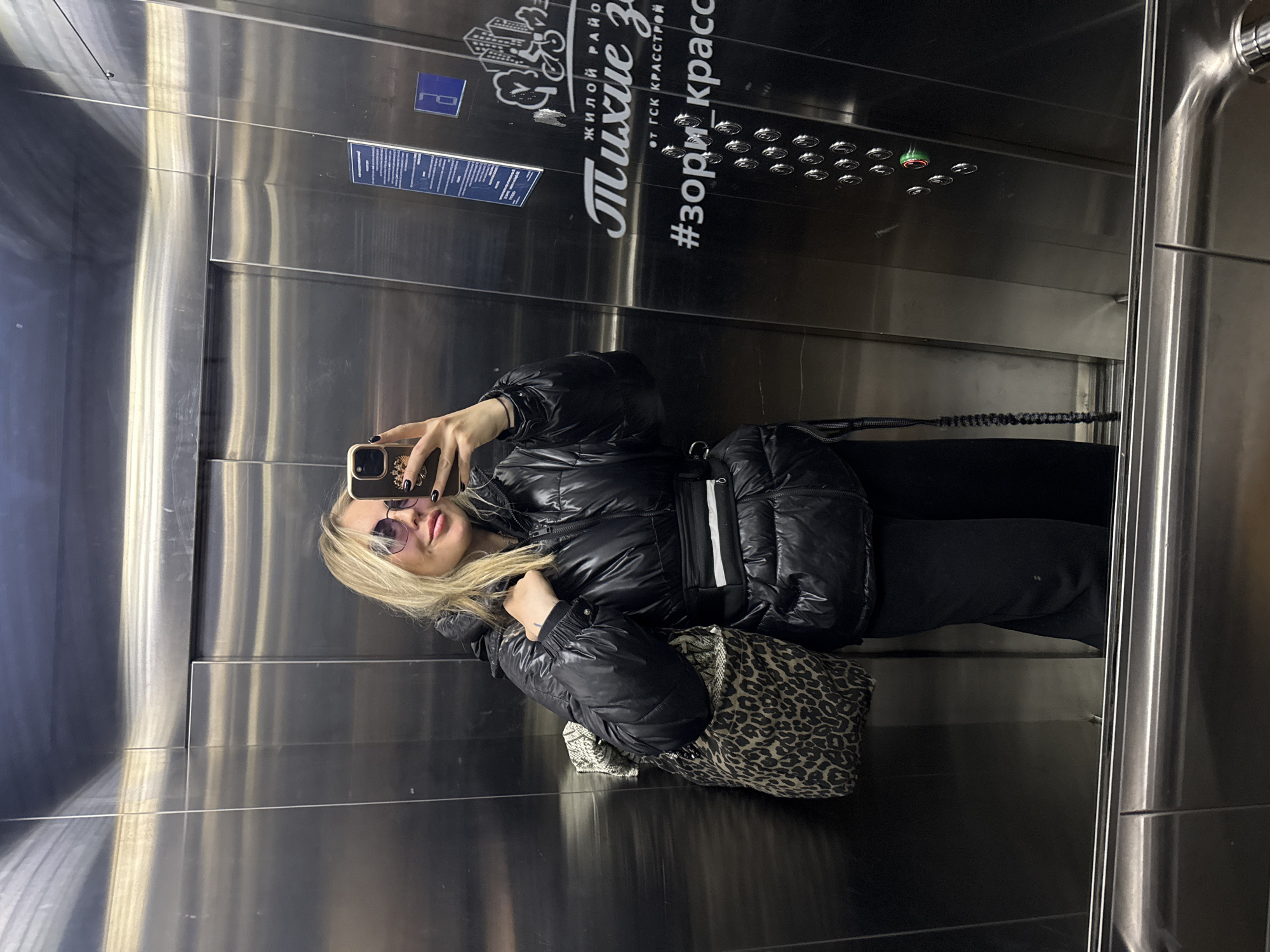 A blonde woman in sunglasses is taking a selfie in an elevator. She is wearing a black puffer jacket and black pants, and carrying a leopard print bag. The elevator has a stainless steel interior. On the elevator wall, there is text in Russian, including "#зори_красск". This hashtag suggests the image was taken in Krasnoyarsk, Russia.