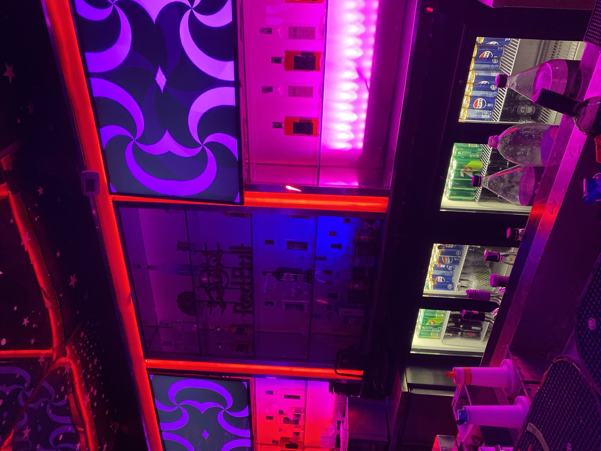 The image depicts the interior of a dimly lit bar, likely in Sharm El Sheikh, Egypt, with vibrant purple and red neon lighting illuminating the scene. In the background, shelves are stocked with various bottles of alcohol, some of which are lit from below with pink and blue lights. Two large screens displaying a purple, abstract pattern are mounted above the shelves. In the foreground, a bar counter is visible, with several bottles and dispensers on its surface. Behind the counter, two refrigerated display cases are filled with cans of soda, including Pepsi, and bottles of water. The overall atmosphere is that of a modern nightclub or bar. There are no people visible in the image.