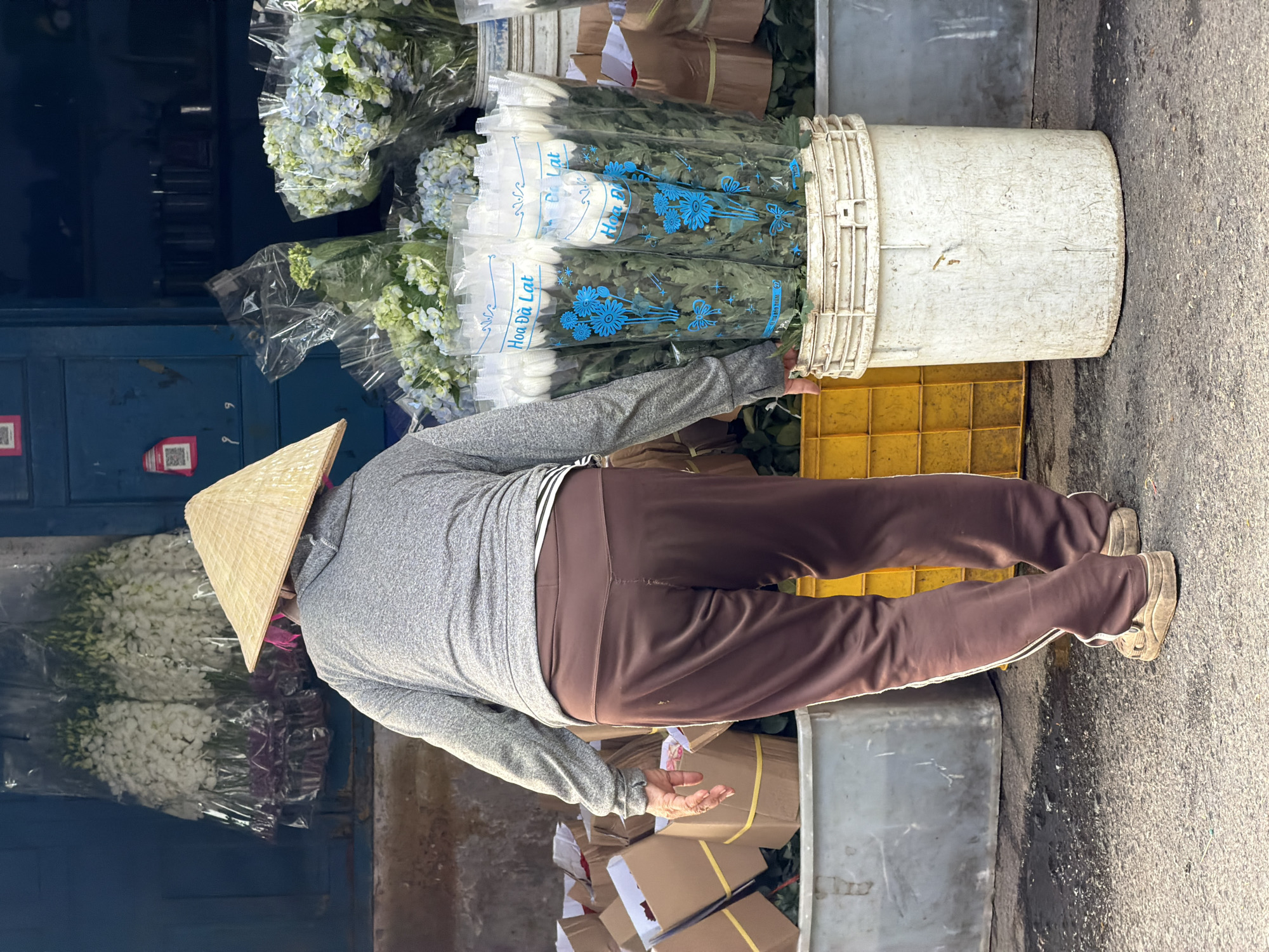 This image depicts an outdoor flower market scene, likely in Nha Trang Ward, Vietnam. A person, seen from behind, is the central subject, wearing a traditional Vietnamese conical straw hat (nón lá), a grey long-sleeved shirt, and dark brown trousers with white side stripes. Their right hand is reaching into a large, white plastic bucket filled with various types of flowers.
The bucket contains numerous bundles of fresh flowers, predominantly light blue and white hydrangeas, and white chrysanthemums, all individually wrapped in clear plastic. The plastic packaging for the chrysanthemums features blue floral designs and clearly displays the Vietnamese text "Hoa Đà Lạt," indicating their origin from Da Lat, a well-known flower-growing region.
The person appears to be engaged in the activity of organizing or retrieving flowers, suggesting a vendor preparing for sale or arranging stock. Surrounding elements include yellow plastic crates on the ground near the bucket, and stacks of cardboard boxes, some tied with yellow string, positioned below a grey metal container on the right. In the background, a dark blue metal cabinet or door is visible, adorned with red QR code stickers, and more wrapped flowers are stacked. The ground is a rough asphalt or concrete surface. The bright, direct sunlight suggests it is daytime, possibly morning, with clear weather.