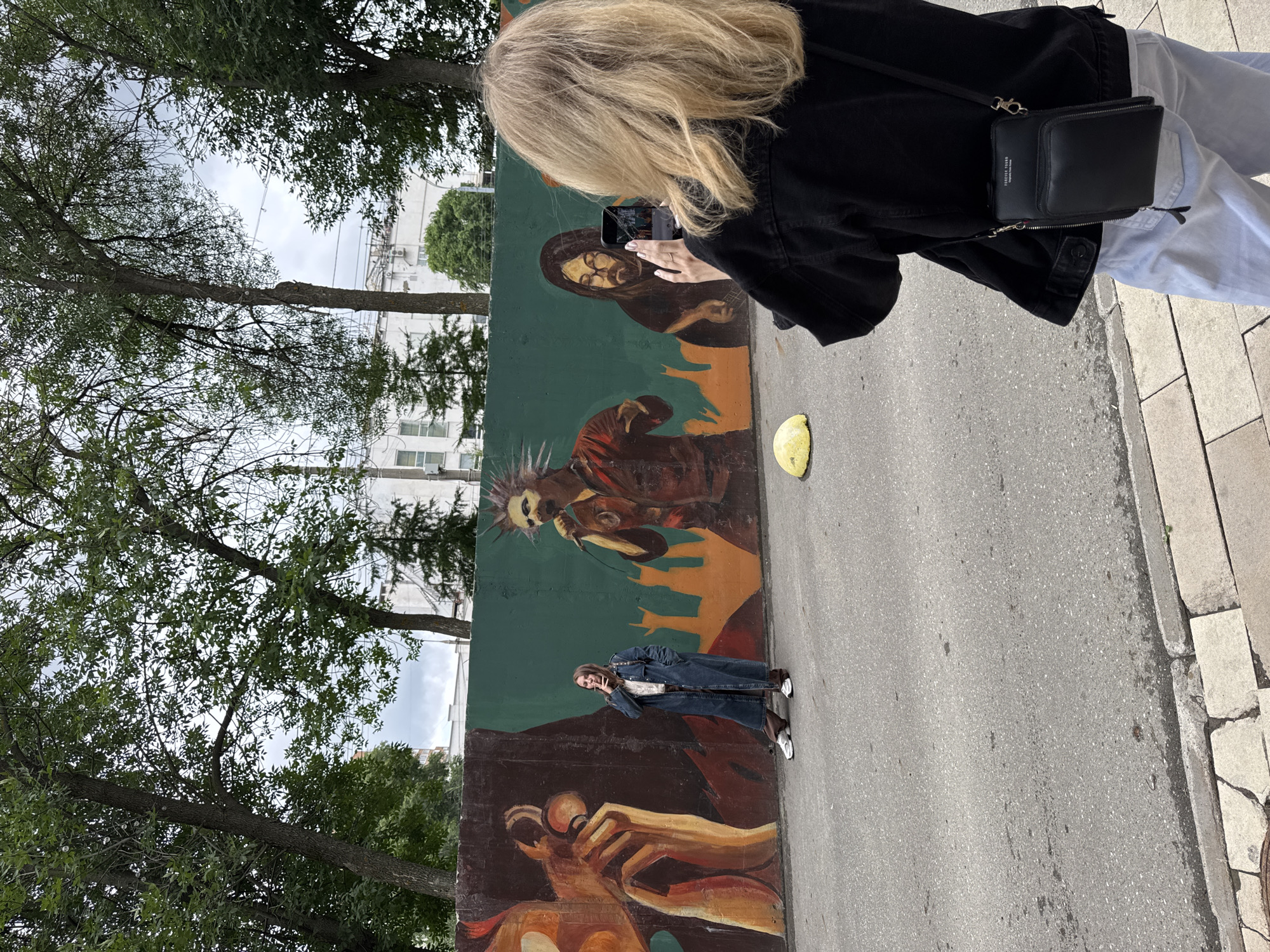 In Izhevsk, Russia, a person with blond hair wearing a black jacket and white pants stands with their back to the camera, holding a phone and taking a photo of a mural on a concrete wall. The mural depicts stylized figures, including one holding a microphone with a mohawk and another playing a guitar. A woman in a long denim coat and dark pants is standing in front of the mural, smoking. The wall is painted with bold colors and abstract shapes. The scene is set on a paved street with a sidewalk. A yellow rounded object is visible on the street near the mural. Trees and buildings can be seen in the background, under a cloudy sky.