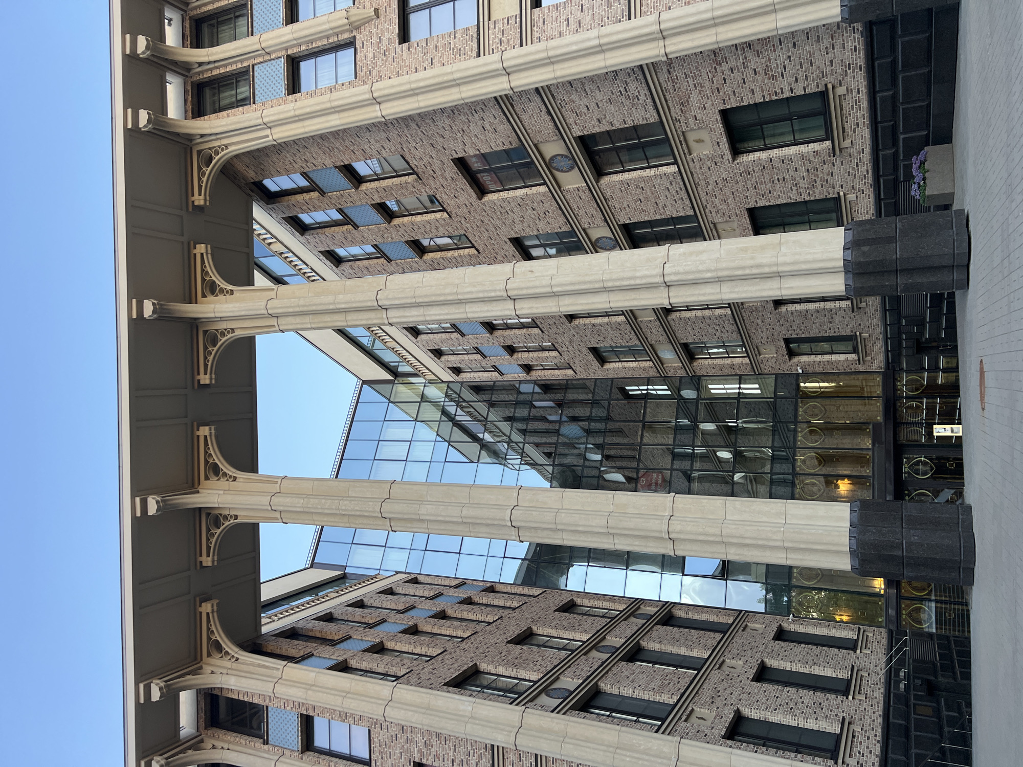 The image shows the exterior of a modern building in Saint Petersburg, Russia, with a clear blue sky above. The building features a facade of light brown brick with dark gray window frames and decorative blue patterned panels between some of the windows. Large, fluted cream-colored columns with ornate archways support a horizontal structure that extends over the entrance area. The central part of the building is a multi-story glass facade, reflecting the sky and the surrounding architecture. The ground level has dark gray paneling and a glass entrance with ornate gold detailing. A planter box with purple flowers is visible to the right of the entrance. The perspective is from a low angle, looking upwards at the building.