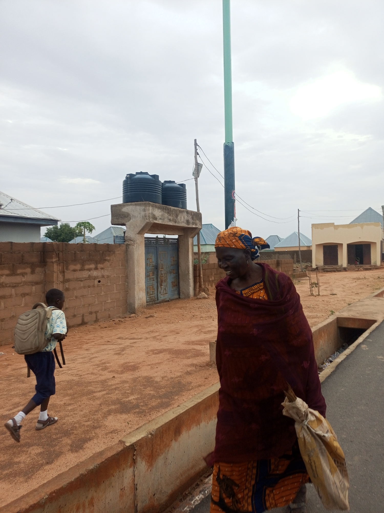 A wide-angle shot captures a street scene in Tempure, Nigeria under a cloudy sky.
An elderly Black woman is walking on the right of the frame. She's dressed in a patterned orange and blue dress. Over it, she wears a burgundy shawl. Her head is wrapped in an orange and blue patterned headscarf. She is smiling, and carries a tattered sack.
To the left, a young Black boy with a backpack is walking away from the viewer on the red dirt. He is wearing sandals and white socks, blue pants, and a light-colored patterned shirt.
Behind them, there is a brick wall that leads to a gated entrance. Atop the entrance are two black water tanks. Beyond the wall and gate, several buildings with tin roofs are visible.
A tall green utility pole is to the left of the woman.
The street has a concrete gutter on the right side, next to the paved road the woman is walking on.