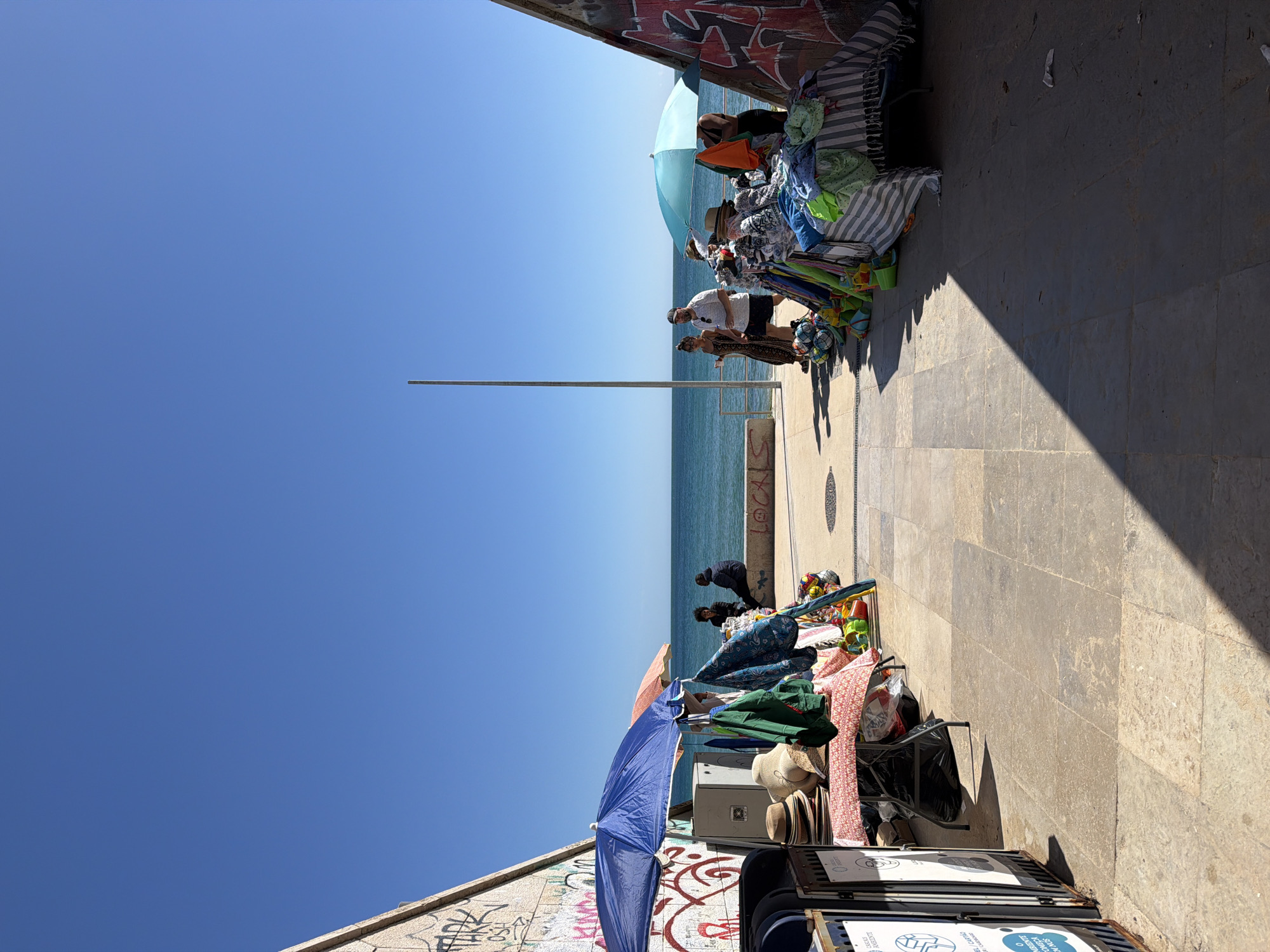 The image captures a street scene in Cascais, Portugal, adjacent to the coastline on a sunny day. People are browsing merchandise laid out on blankets and tables. Umbrellas provide shade for the vendors. The merchandise appears to be a variety of items, including clothing, souvenirs, and beach accessories. The backdrop includes the ocean and a clear blue sky. Street art is visible on a wall. Street refuse containers are present in the foreground. The photograph is taken from a ground-level perspective, facing the sea. A solitary pole rises vertically into the clear sky.