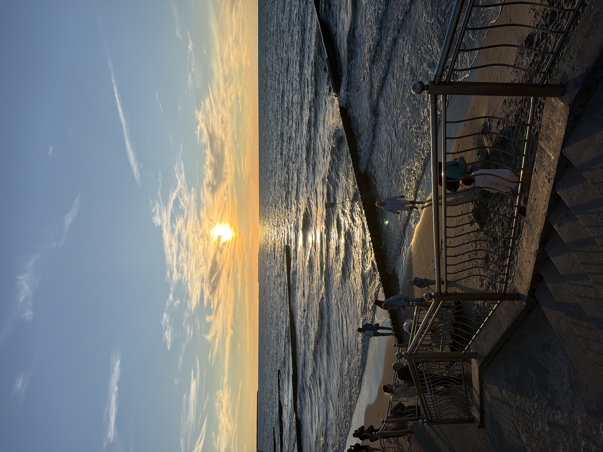 A vertical, eye-level shot captures a sunset over the Baltic Sea from the promenade in Zelenogradsk, Russia. The sun, a bright orb, is positioned low on the horizon, casting a golden, shimmering reflection across the choppy waves. Scattered white clouds streak across the clear blue sky, tinged with orange and yellow near the sunset.
In the foreground, a concrete staircase descends to the sandy beach, bordered by a metal railing with decorative scrolls. Several individuals are visible on the promenade and the beach. Some are walking, while others appear to be conversing. A few people are near the water's edge, and one person is wading in the sea. Wooden groynes, partially submerged, break the rhythm of the waves extending out into the water. The scene is bathed in the warm, soft light of the setting sun.