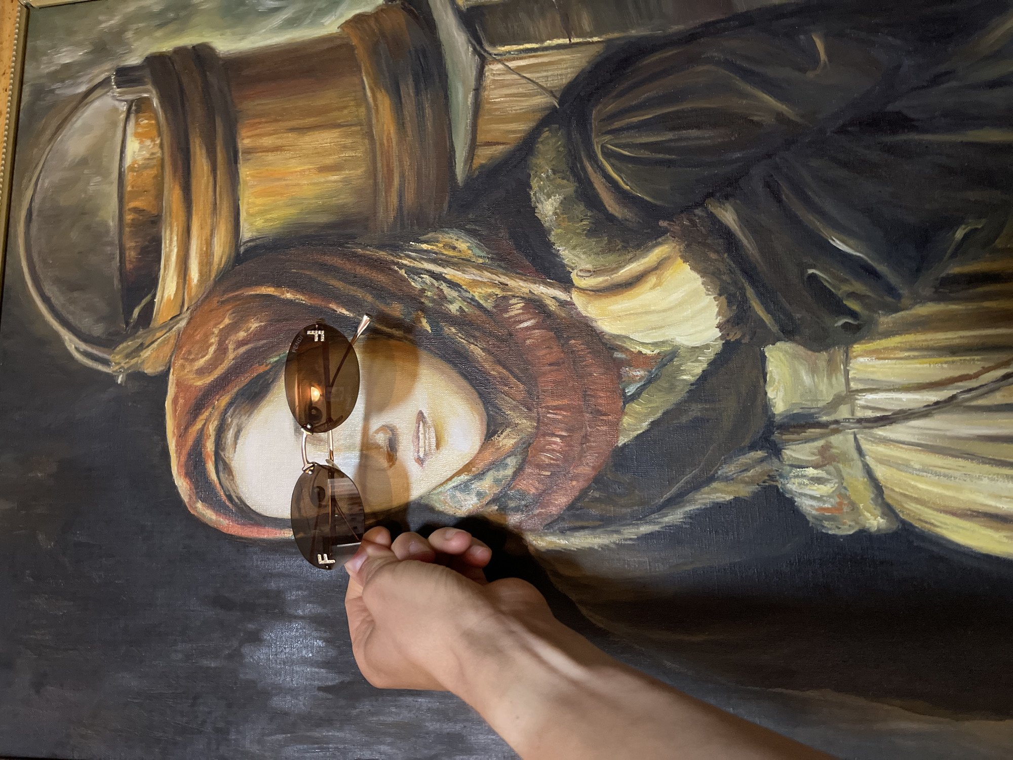 A painting of a woman with cargo on her head is visible, held by a hand with sunglasses in front of the woman’s face. The painting is likely located in Перевозное, Russia. The woman in the painting is wearing a head covering and appears to be carrying bundles or containers on her head, possibly related to trade or transportation. The overall style and colors suggest an oil painting.