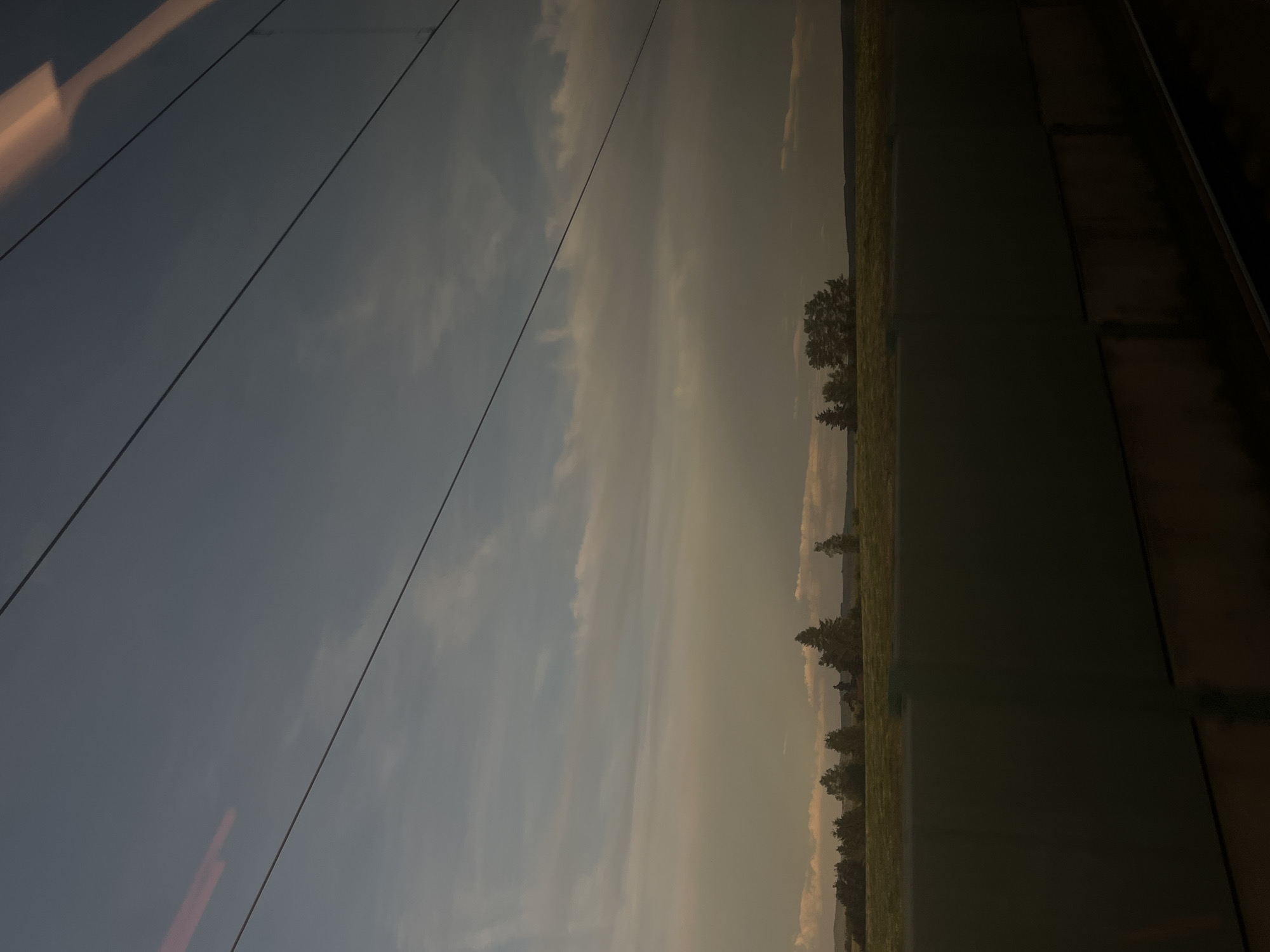 The image captures a view from a train window, looking out at a rural landscape on the outskirts of Ilmenau, Germany. The sky is a gradient of dark blue at the top, transitioning to lighter shades with wispy clouds, and a warmer, hazy tone near the horizon. Several dark, straight lines, likely overhead power lines for the train, traverse the sky. Below the sky is a band of green land, dotted with clusters of trees, some appearing as dark silhouettes. In the foreground, a dark, solid barrier, possibly part of the train or an overpass, obscures the lower portion of the scene. The overall impression is one of a moving perspective, capturing a fleeting moment of the countryside.