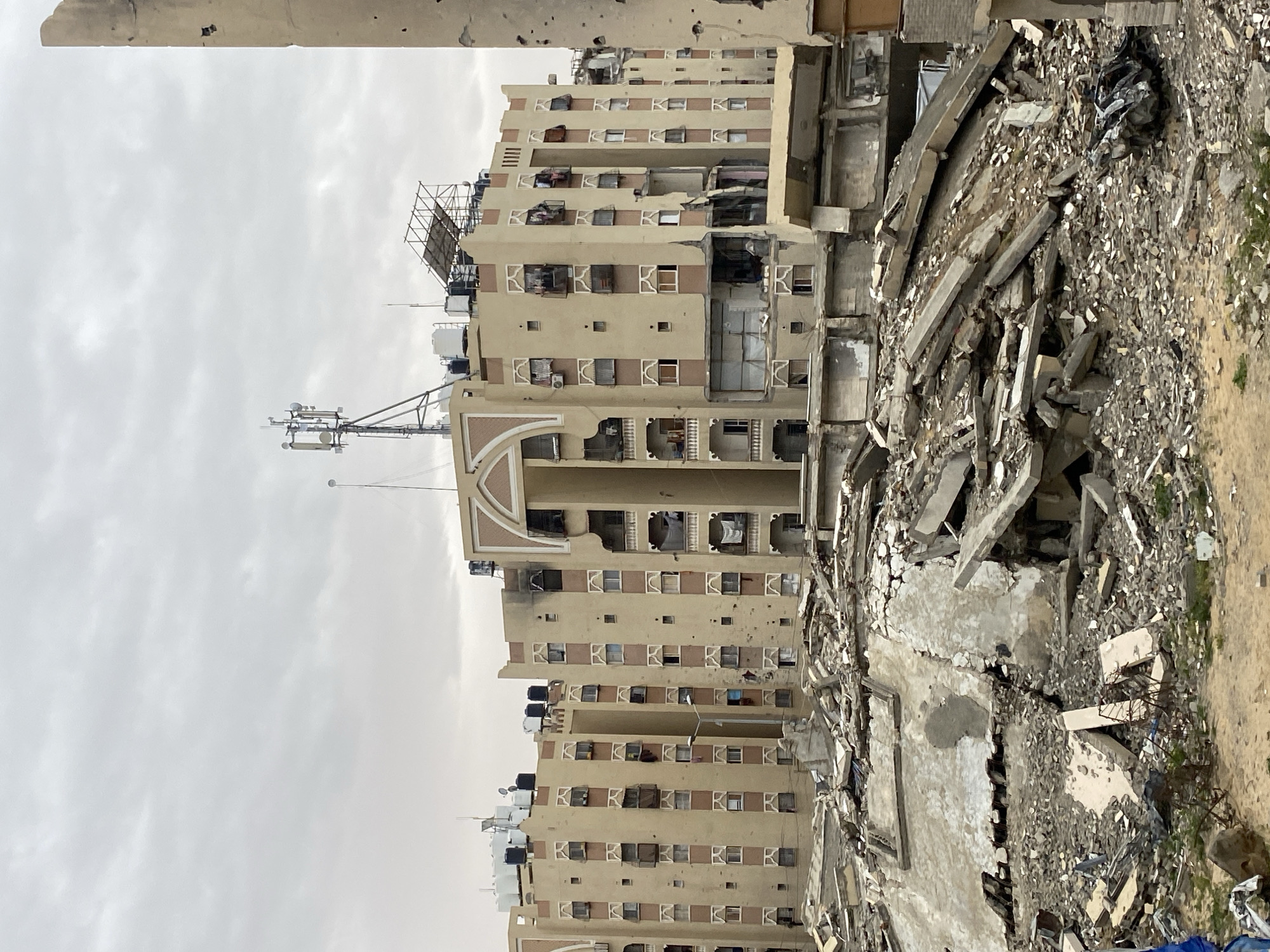 The image shows a building in Khan Yunis, Palestinian Territory, severely damaged by what appears to be conflict. A multi-story beige building stands partially intact, with visible damage including holes and missing sections. Adjacent to the building is a large pile of rubble and debris, indicating a collapsed structure. The sky is overcast. There are no visible people. Antennas and water tanks are on top of the building.