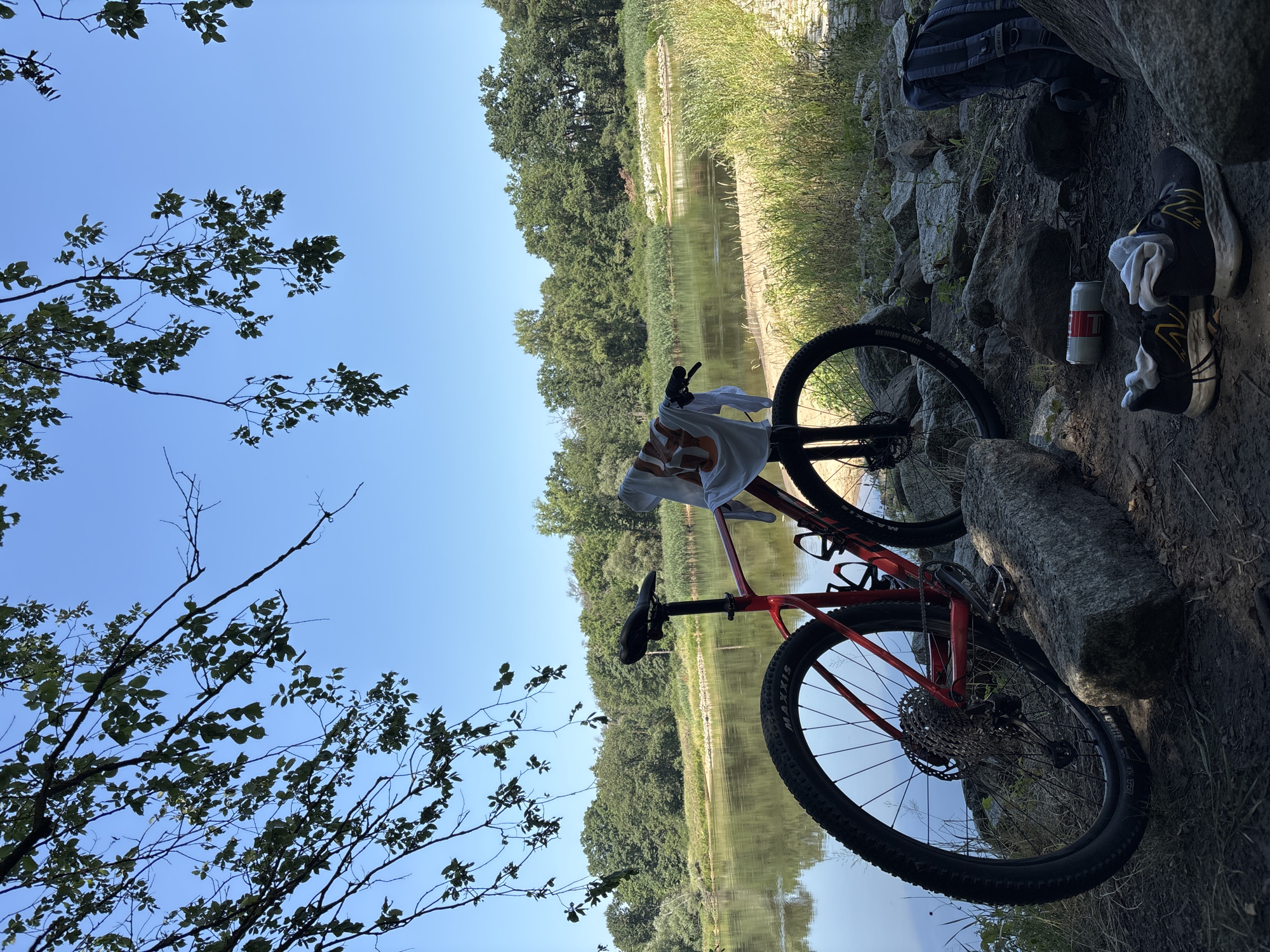 The image depicts a scene near a body of water, likely a river or lake, bordered by trees and vegetation under a clear blue sky. In the foreground, a red bicycle is parked next to a large rock. A white t-shirt with an orange graphic is draped over the handlebars. On the ground near the bicycle, a pair of black and yellow athletic shoes with white socks are placed, alongside a metallic can and a dark blue backpack. The terrain is rocky and covered with dirt and grass. The overall atmosphere suggests an outdoor recreational setting. Without additional information or context, the specific location in Zielona Góra, Poland cannot be determined from the image itself.