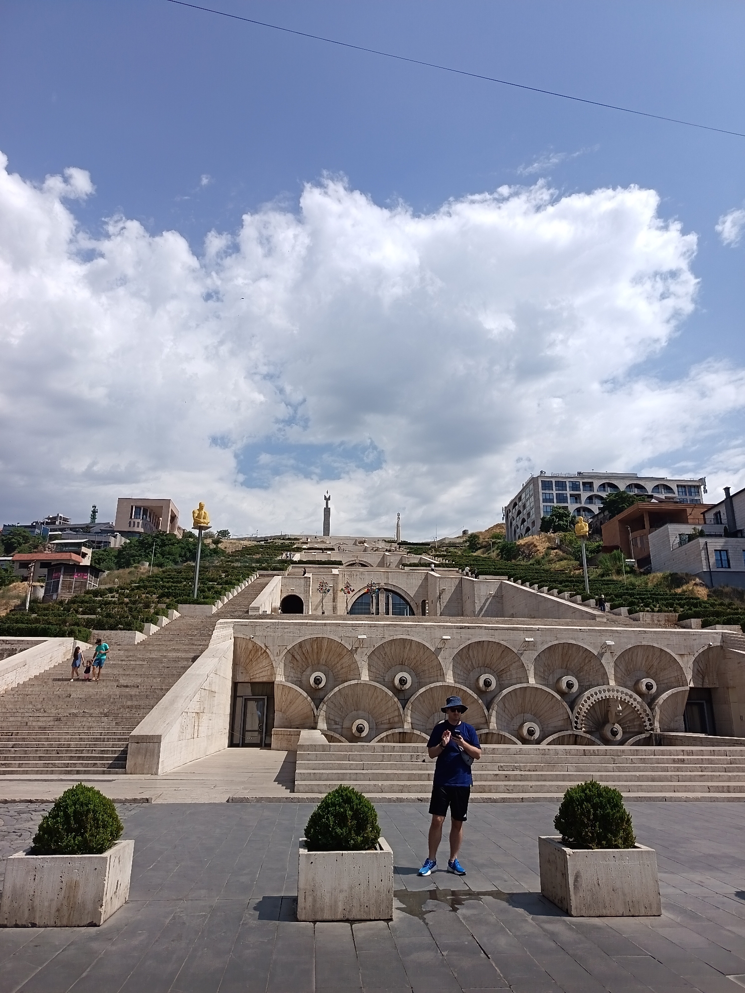 A full outdoor shot captures a man in a blue t-shirt, black shorts, a blue hat, and blue sneakers, standing in a paved area in front of a large, tiered stone structure. The man is looking down at his phone and holding it in his right hand. Two large, square planters with green bushes flank him, one on his left and one on his right. The stone structure behind him features multiple levels of stairs and decorative arches with circular elements. In the background, a wide staircase leads up a hillside dotted with greenery and buildings. The sky above is blue with scattered white clouds, and a thin black wire is visible across the top of the frame.