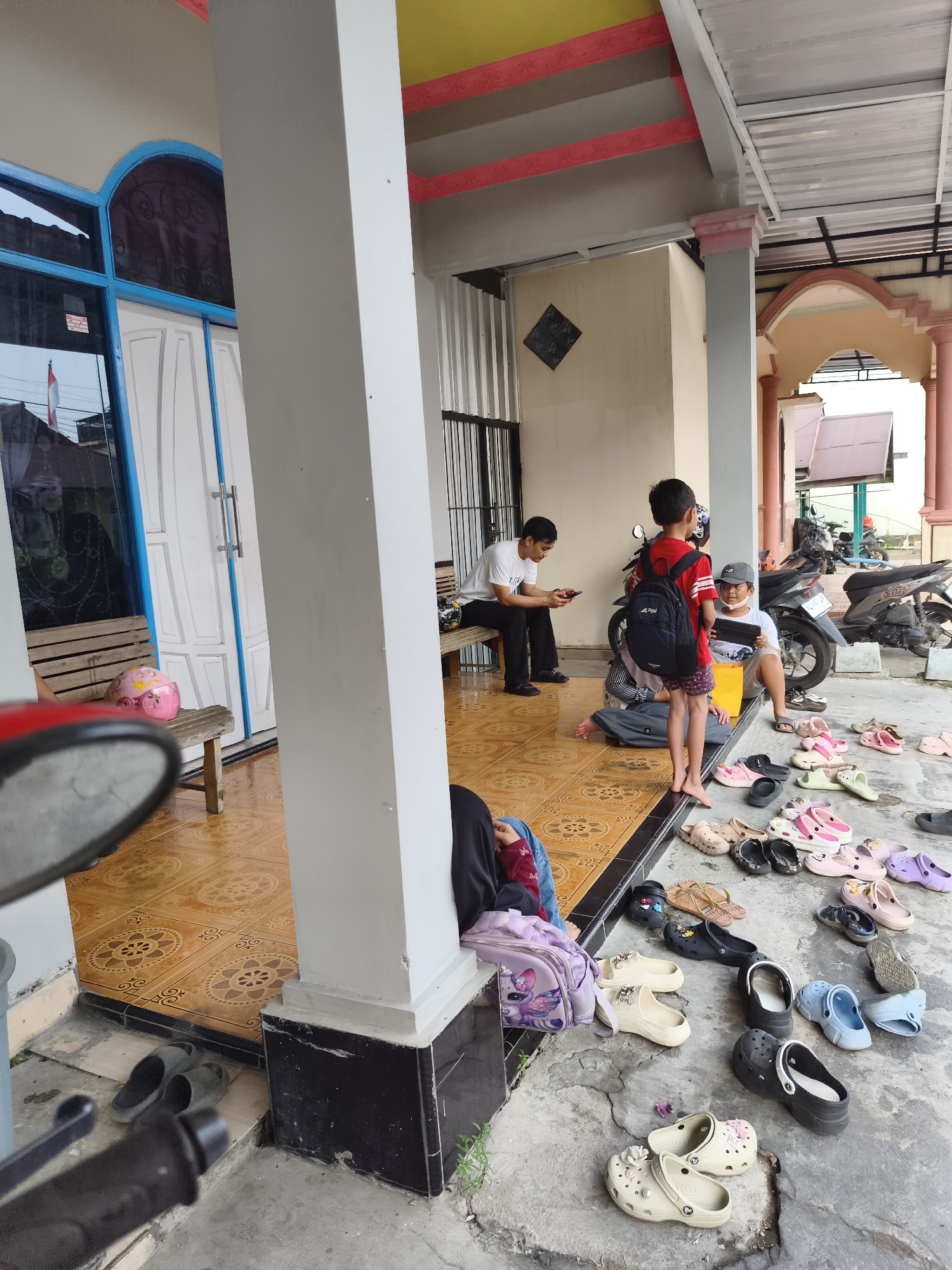 The image depicts an outdoor scene, likely a residential porch in Tanjung Redeb, Indonesia, given the style of architecture and the presence of motorcycles commonly seen in Indonesian households. Several people, including children and adults, are present. One adult male is seated on a wooden bench, looking at his phone. Two children are standing, one in a red shirt and black shorts holding a backpack, and another in a white shirt and gray cap, holding a tablet. Several other individuals are seated or lying on the floor, some with backpacks. A variety of footwear, predominantly sandals and flip-flops, are scattered on the tiled porch floor. Motorcycles are parked in the background. The environment appears to be a covered outdoor space with tiled flooring and a white pillar supporting the roof. The overall impression is of a casual gathering or waiting area.