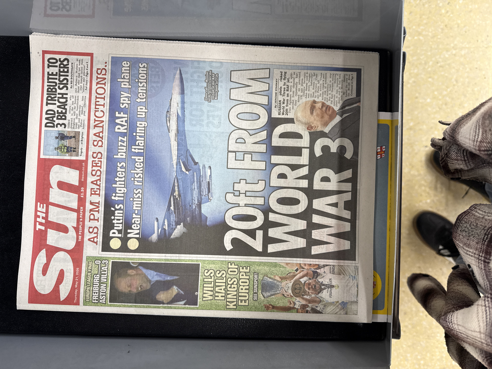 The image shows a copy of "The Sun" newspaper, dated Thursday, May 21, 2026, lying in what appears to be a display case. The main headlines are "20ft FROM WORLD WAR 3," related to Russian fighters buzzing an RAF spy plane, and "AS PM EASES SANCTIONS." Additional headlines include "DAD TRIBUTE TO 3 BEACH SISTERS," and "WILLS HAILS KINGS OF EUROPE." The front page also features the score of a Europa League final between Freiburg and Aston Villa. Part of a person's legs, wearing black shoes and a patterned coat, is visible to the right of the newspaper.
There is no information about Great Yarmouth in the picture.