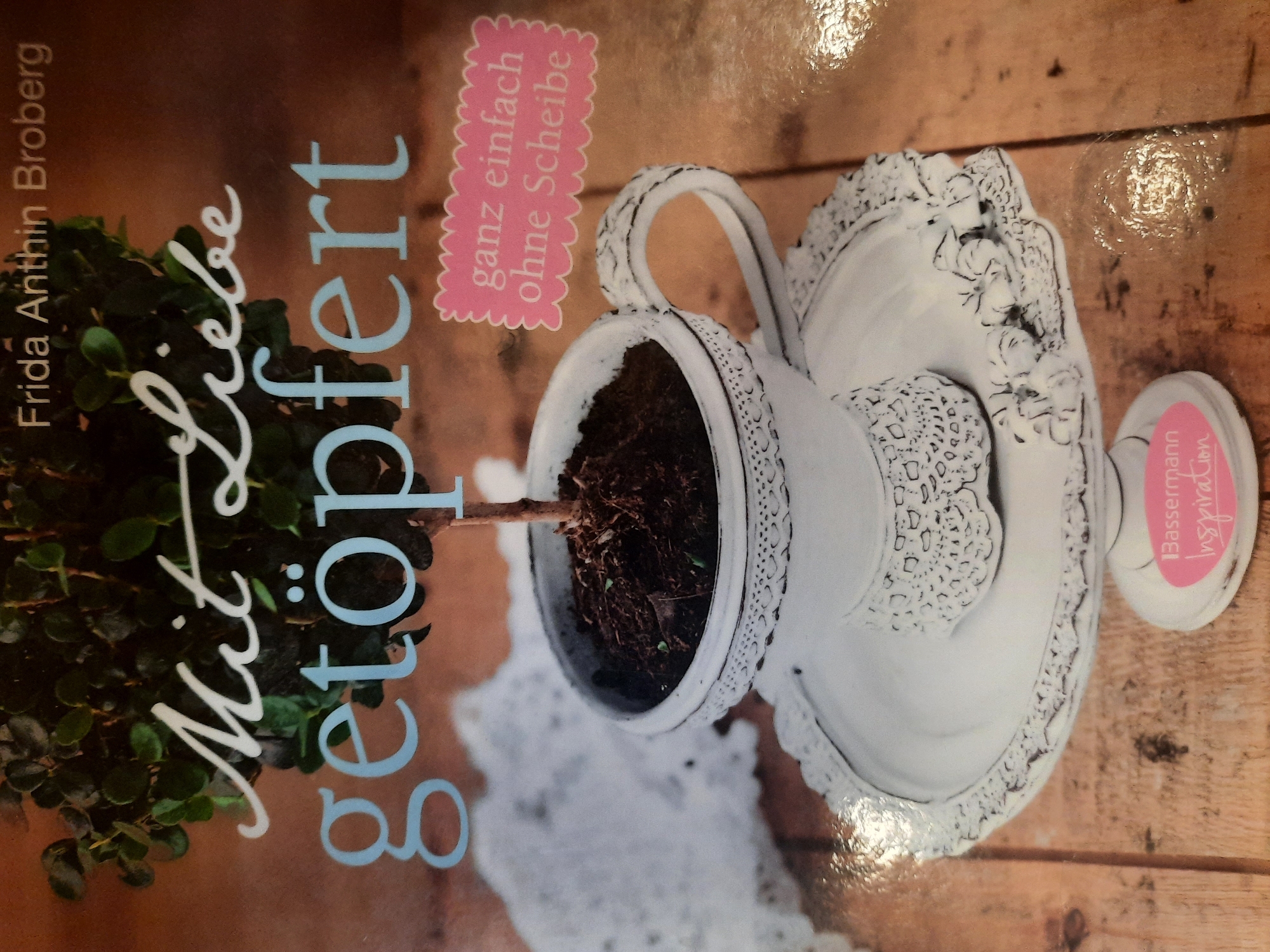 The image is a close-up of a book cover with the title "Mit Liebe getopfert" (Potted with Love) and the author "Frida Anthin Broberg". The cover features a decorative white teacup placed on a white cake stand, both adorned with intricate lace-like detailing. The teacup contains dark soil and a small green plant. To the right of the teacup, a pink tag with scalloped edges contains the German text "ganz einfach ohne Scheibe" (very simple without glass). At the bottom of the cake stand, a pink oval label reads "Bassermann Inspiration". The background is a wooden surface. The image does not contain any information about the city of Rosenheim, Germany.
