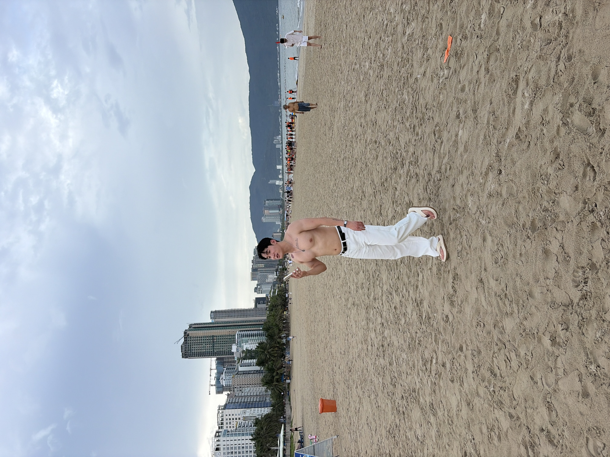 A shirtless man with tattoos on his chest walks on a sandy beach in Da Nang, Vietnam, holding a smartphone. He is wearing white pants and flip-flops. The background shows a line of high-rise buildings, palm trees, and a cloudy sky. In the distance, a group of people are gathered near the water, some wearing orange life vests. The beach is sparsely populated with a few other individuals walking or standing. An orange trash can is visible on the left side of the frame.