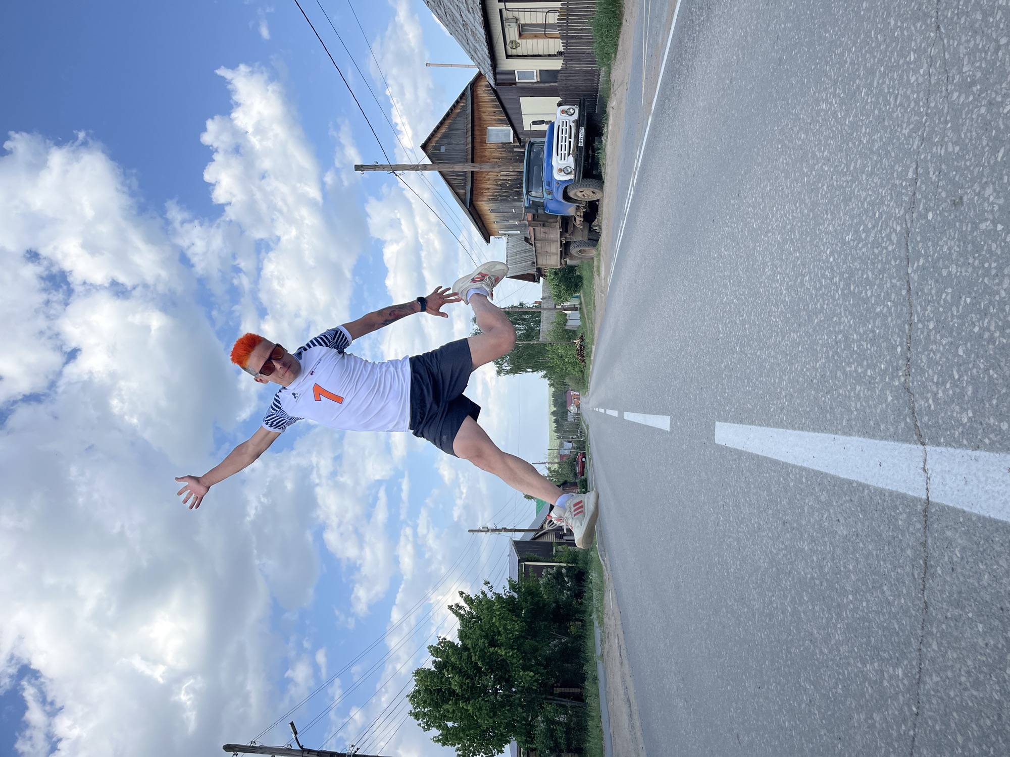 A man with orange hair and sunglasses, wearing a white sports shirt with the number "1" on it and black shorts, is suspended horizontally in the air, appearing to be parallel to a road with a white dividing line. He is outstretched, with one arm pointing towards the sky and the other towards the ground. His legs are also extended. A blue truck is parked next to a wooden house. The sky is mostly blue with white clouds. There are utility poles along the roadside. The scene is set in the city of Chaikovsky, Russia, as indicated by the license plate on the truck.