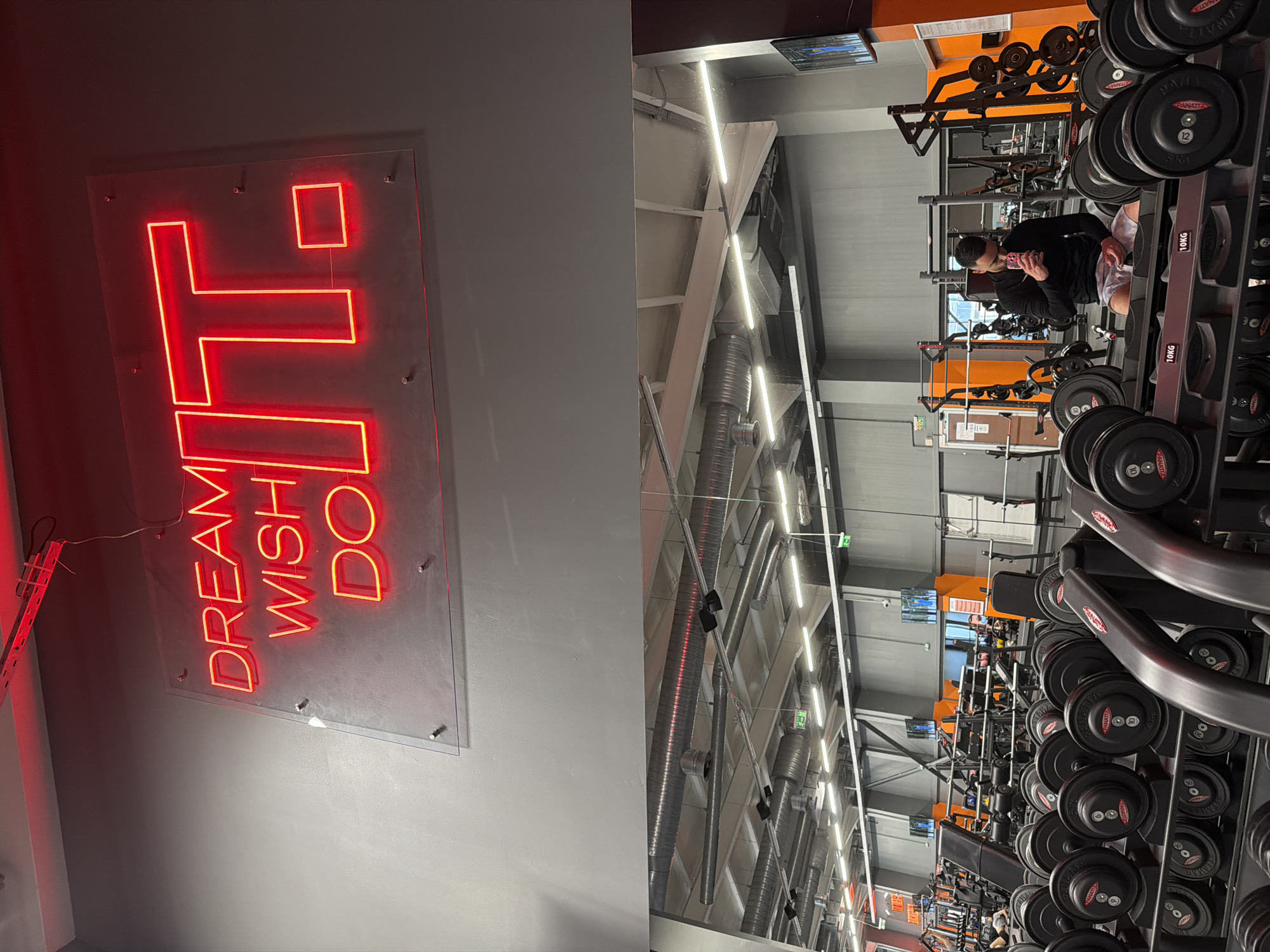 The image shows an interior shot of a gym. On the left, a neon sign with the words "DREAM WISH DO IT" is mounted on a gray wall. On the right, rows of weights are visible, and a man in a black long-sleeved shirt and patterned shorts is sitting on a weight rack, looking at his phone. The gym is well-lit with overhead lights, and the architecture includes metal beams and supports. The gym appears to be located in Plovdiv, Bulgaria.