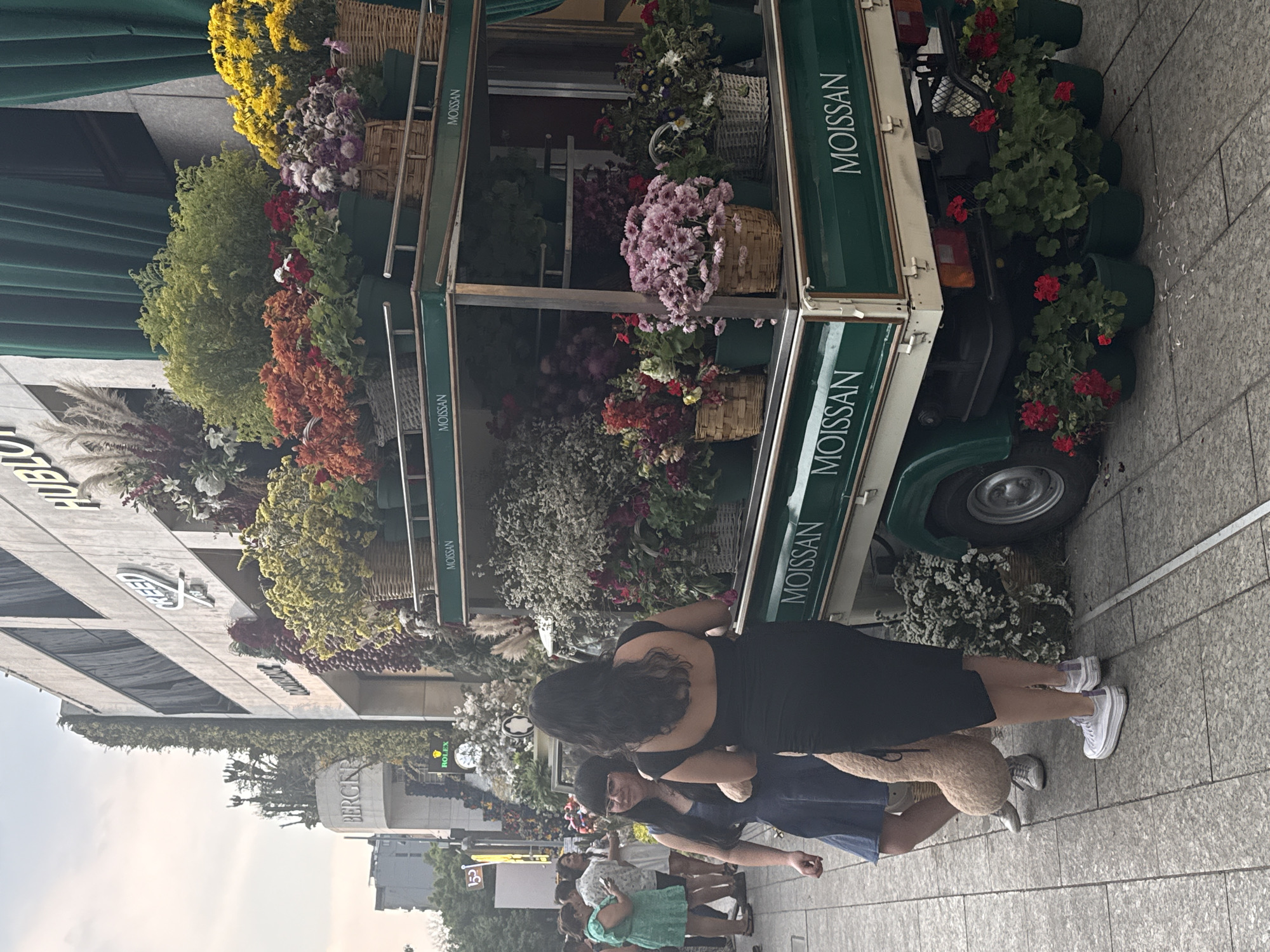 The image shows a group of people on a sidewalk in Mexico City, Mexico. In the background is a building with the word "BERGKG" on it and a Rolex clock. There is also a flower-covered truck with the word "MOISSAN" on it. The people are standing or walking on a sidewalk. A woman in a black dress is hugging another person. Several other people are nearby. The environment is urban, with buildings and greenery visible.