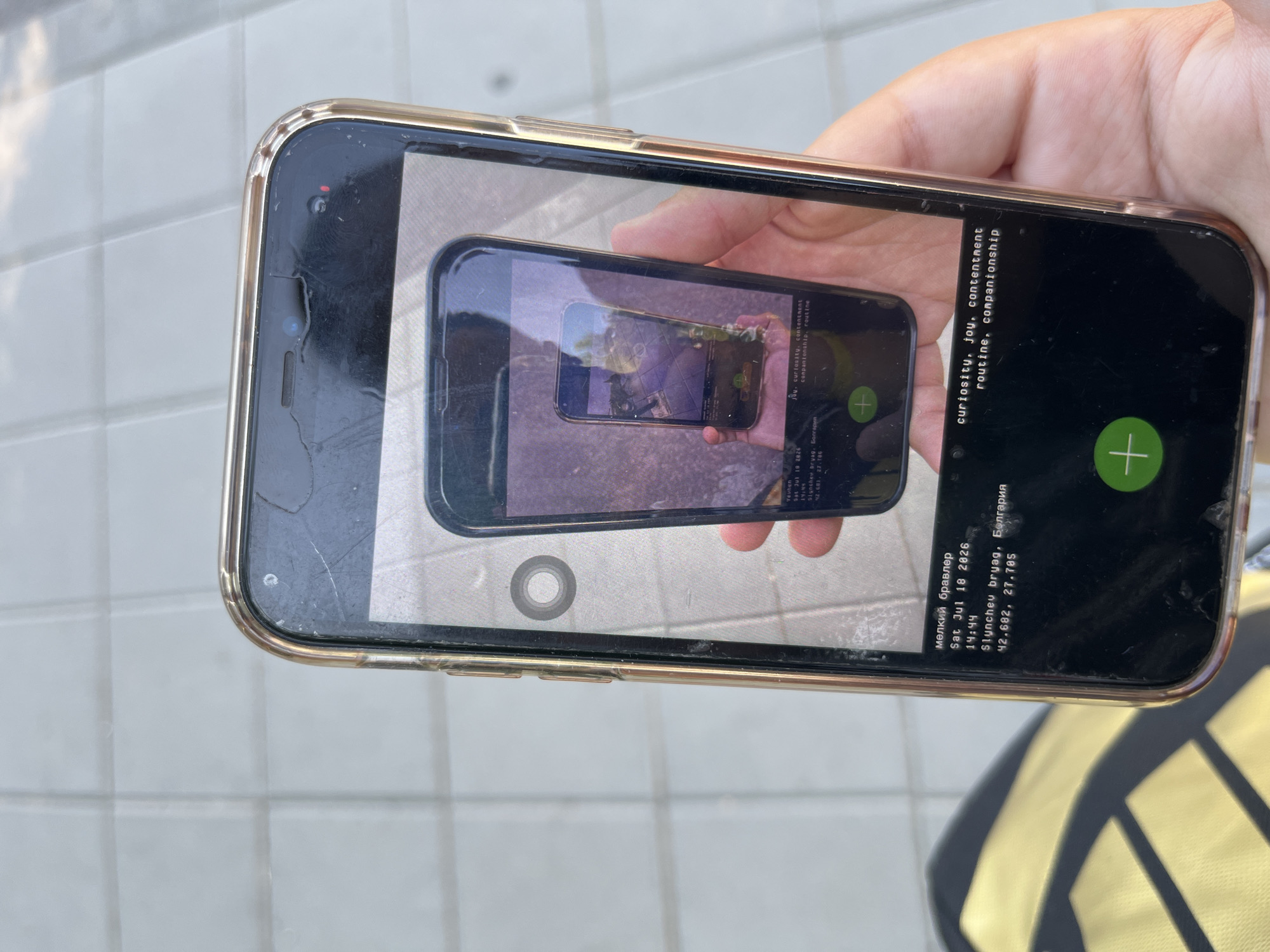 A person is holding a smartphone with a clear case, displaying a photograph of another smartphone held by a hand. This second smartphone shows a reflection of the first smartphone, creating a recursive image. The displayed photograph on the phone contains text overlays. The text includes what appears to be a date and time: "Sat Jul 18 2026 14:44," followed by a location: "Slynchev bryag, Bulgaria 42.682, 27.705." Another line of text reads "meлкий бравелр". A further set of text overlays on the secondary phone's screen reads "curiosity, joy, contentment routine, companionship". The overall scene is a close-up shot of the hand holding the phone, with the background out of focus and appearing to be an outdoor paved surface.